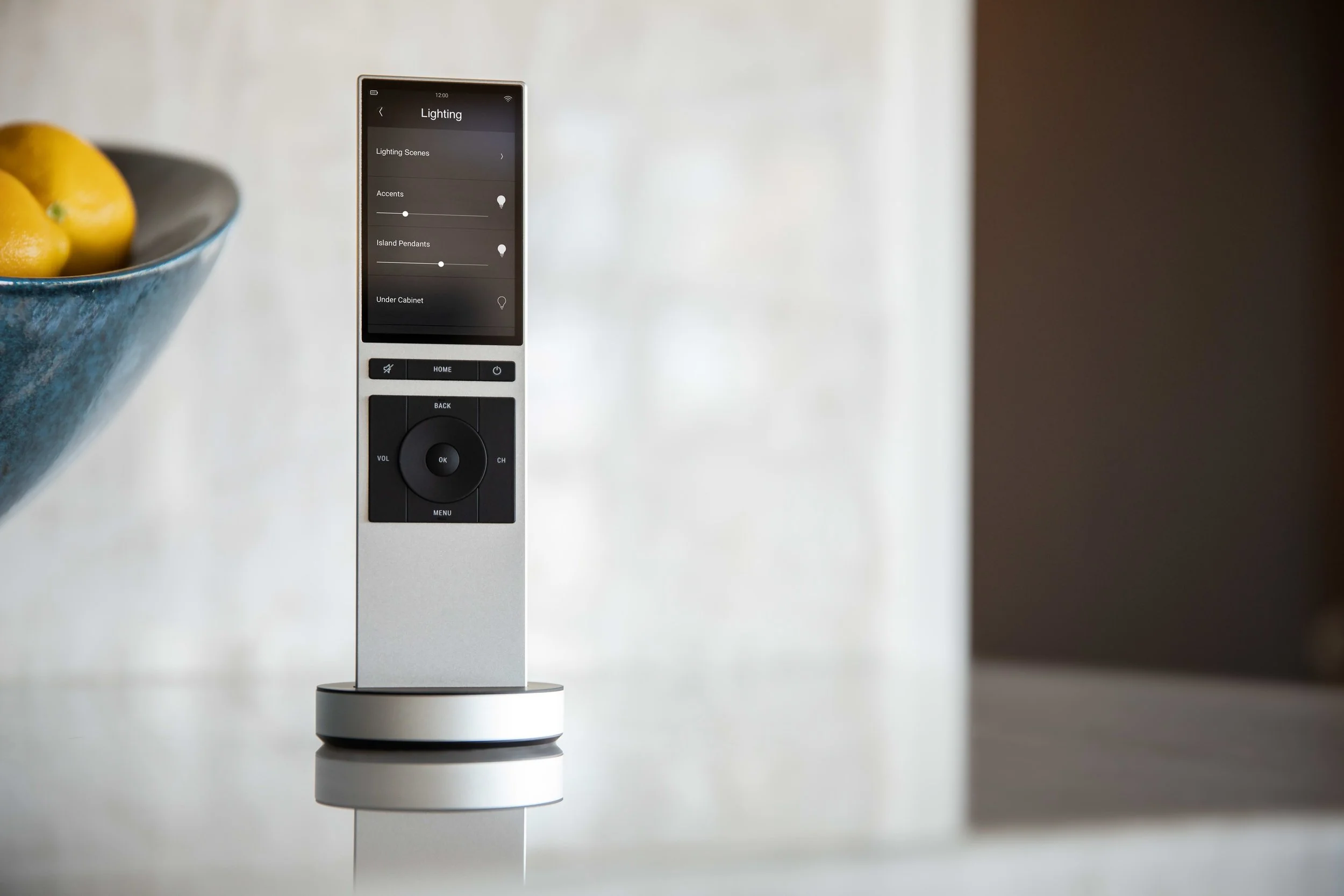 A remote control with a digital screen displaying lighting settings, placed upright on a dock on a white surface with a blurred background.