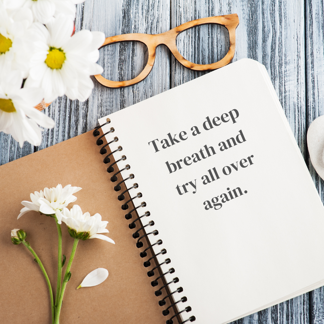 Open spiral notebook with the quote, "Take a deep breath and try all over again," on a wooden surface surrounded by white daisies, a pair of wooden glasses, and a small round stone.