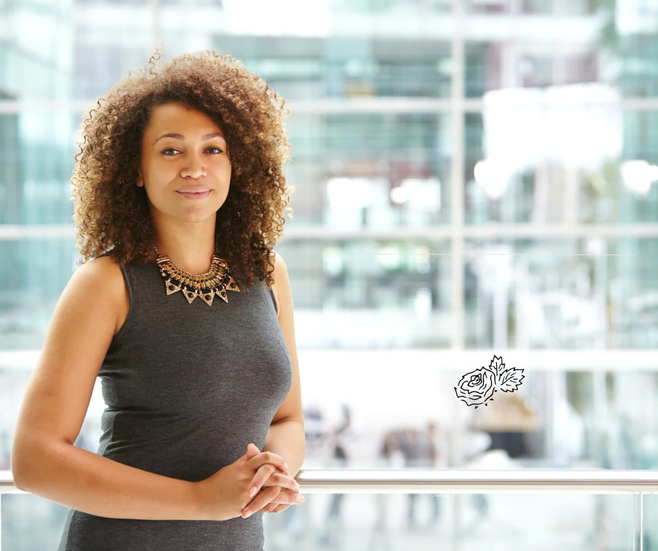 A woman with curly hair wearing a sleeveless dark gray dress and a statement necklace stands in front of large windows in a modern building, with a floral line drawing near the bottom right of the image.