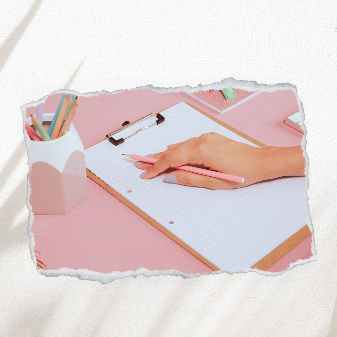 A person's hand holding a pink pencil over a lined notebook on a pink desk, with a white container of colored pencils nearby.