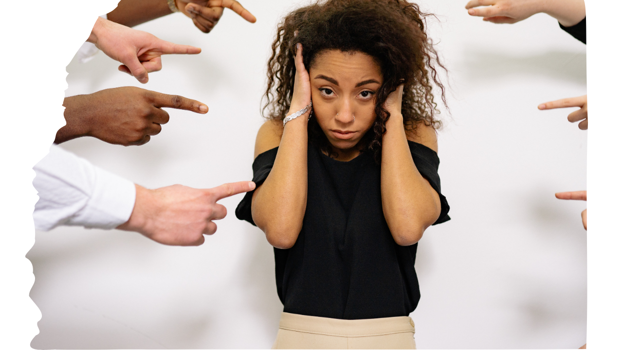 A woman with curly hair looks stressed with her hands on her head, surrounded by multiple hands pointing fingers at her.
