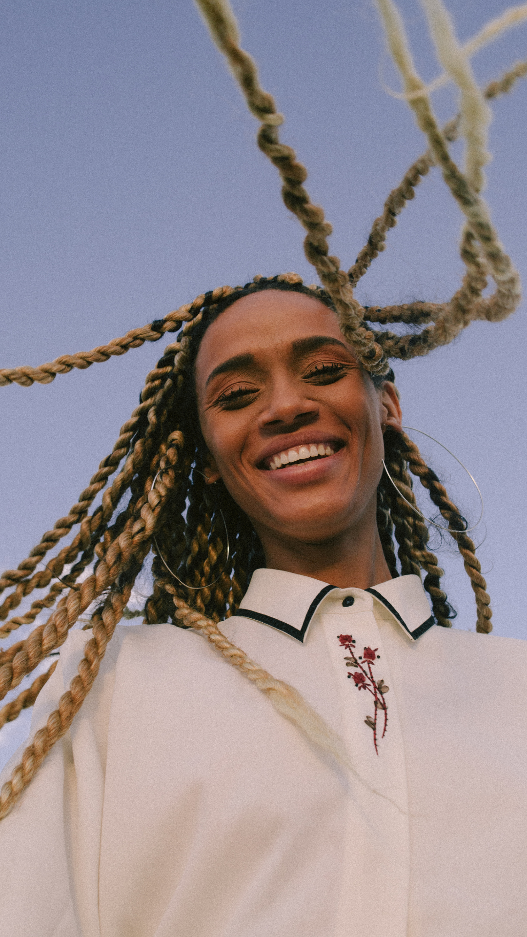 A smiling woman with blonde braids and hoop earrings, looking down at the camera, wearing a cream-colored shirt with embroidered red flowers, against a blue sky background.