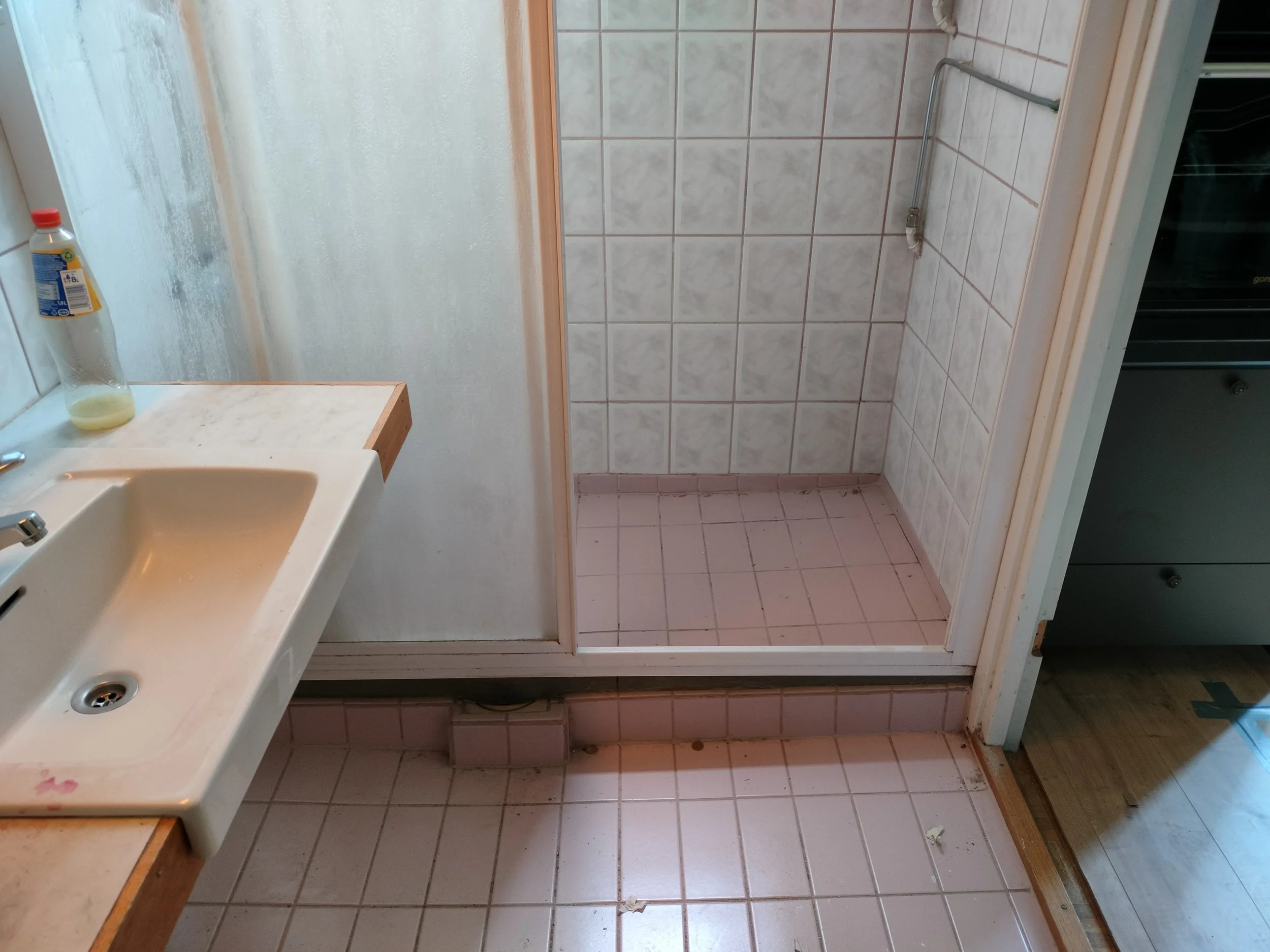 A small bathroom with a sink on the left, a frosted glass shower door at the back, and a tiled shower area with white and pink tiles. There is a bottle on the sink and some debris on the floor.