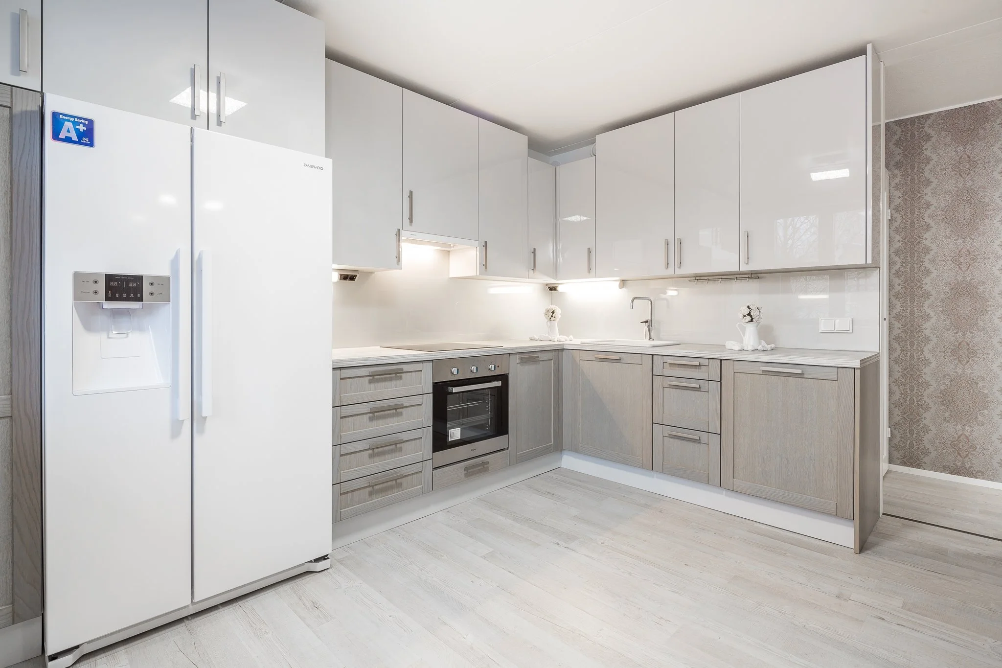 Modern kitchen with white upper cabinets, light gray lower cabinets, a white refrigerator with water and ice dispenser, and stainless steel oven, all in a light-colored room.