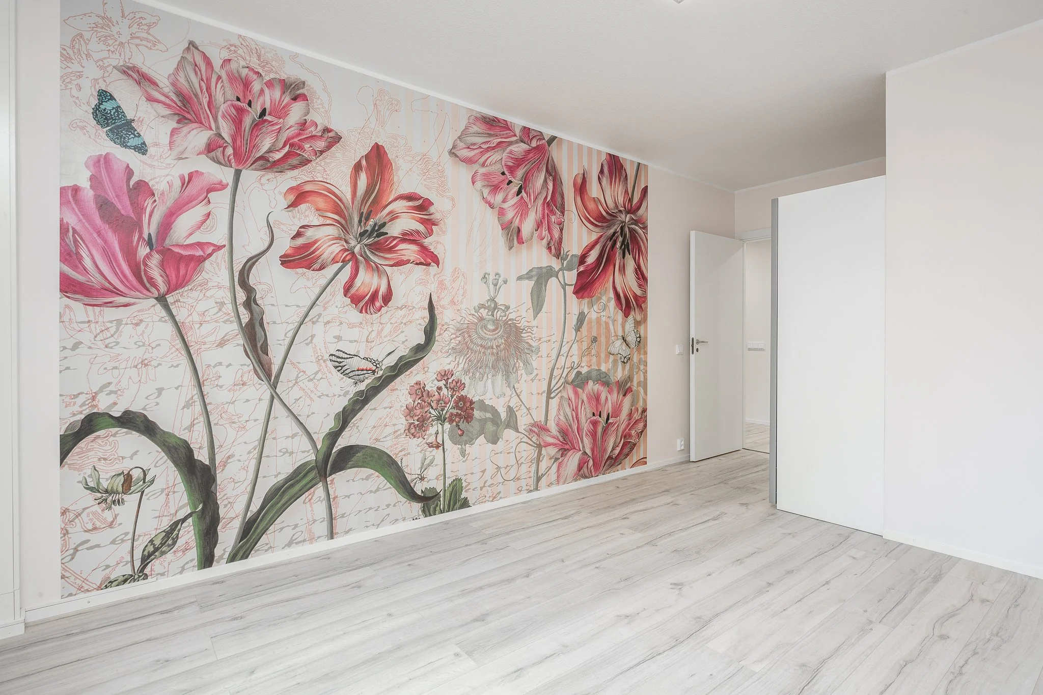 Empty room with floral wallpaper featuring large pink flowers, butterflies, and butterflies on a cream background, wood flooring, open white door.