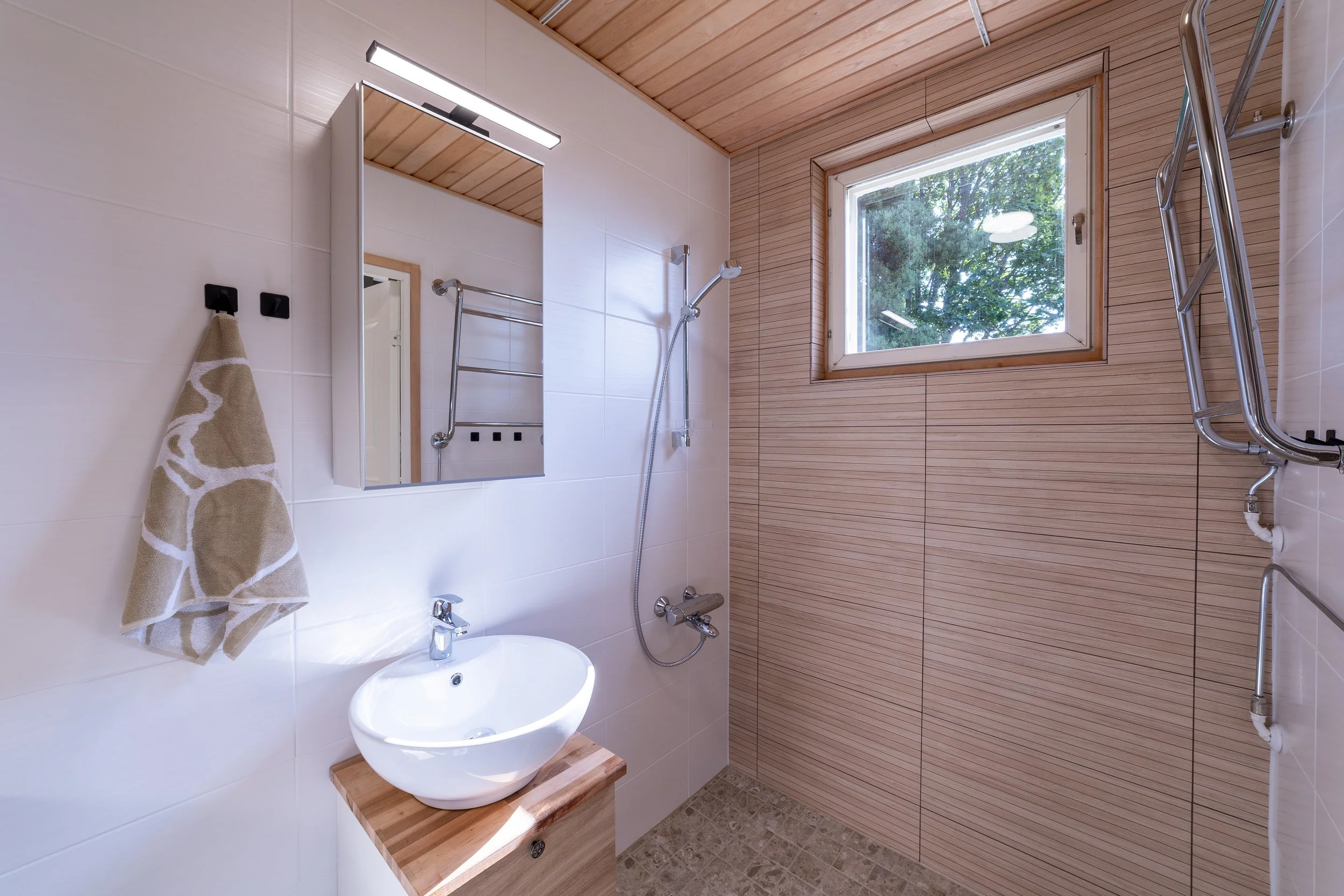 Bathroom with wood ceiling and walls, white tiled walls, a mirror, a towel rack with a towel, a small sink, a shower head, and a window showing trees outside.