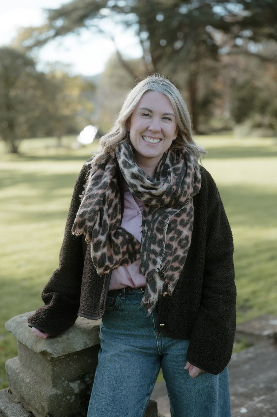 A woman with blonde hair smiling outdoors, wearing a black jacket, a leopard print scarf, a pink blouse, and blue jeans, standing next to a stone bench in a park with trees and grass.