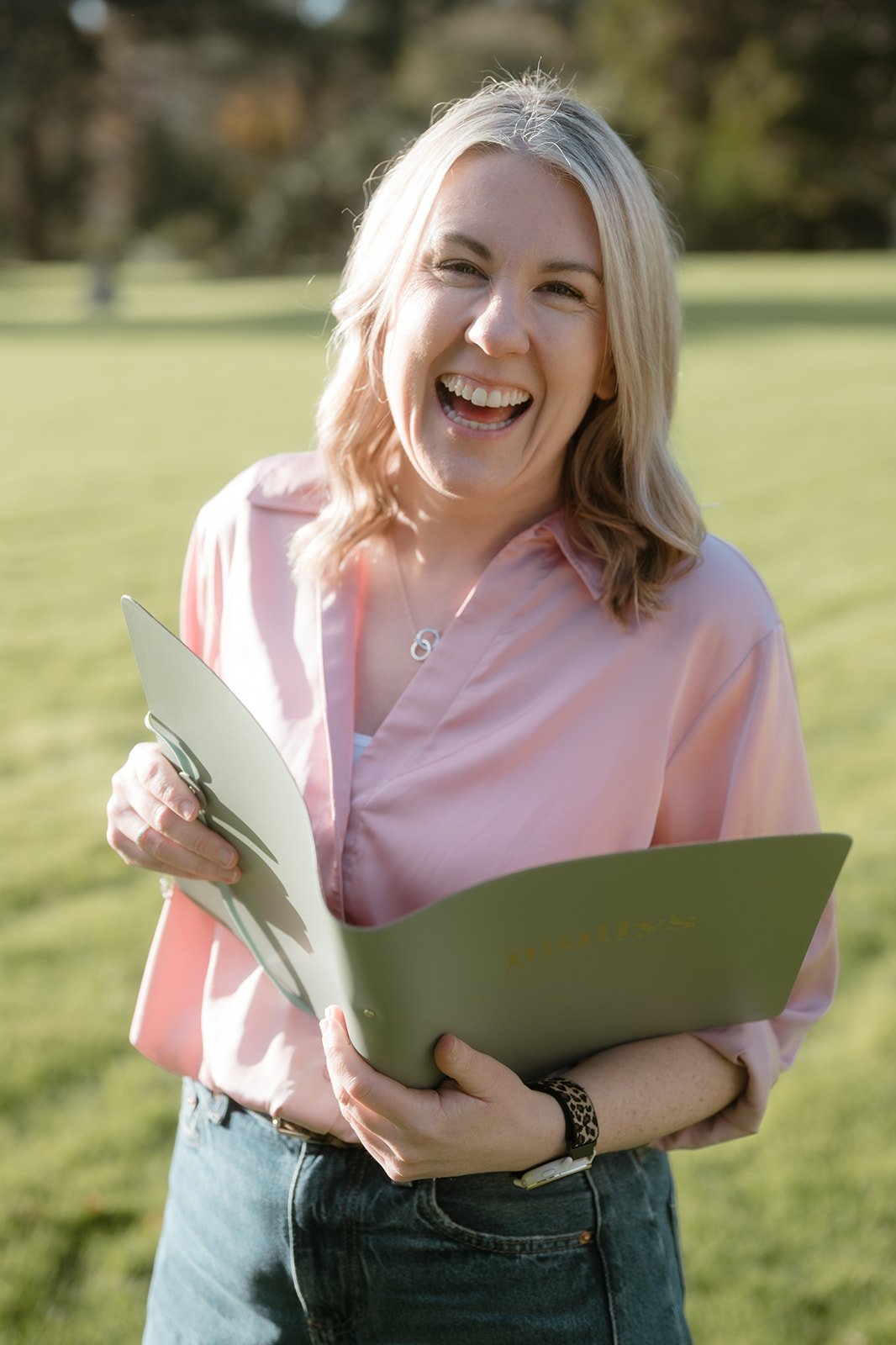 A woman with blonde hair wearing a pink shirt and dark jeans, smiling and holding a green folder outdoors in a park with green grass and trees in the background.