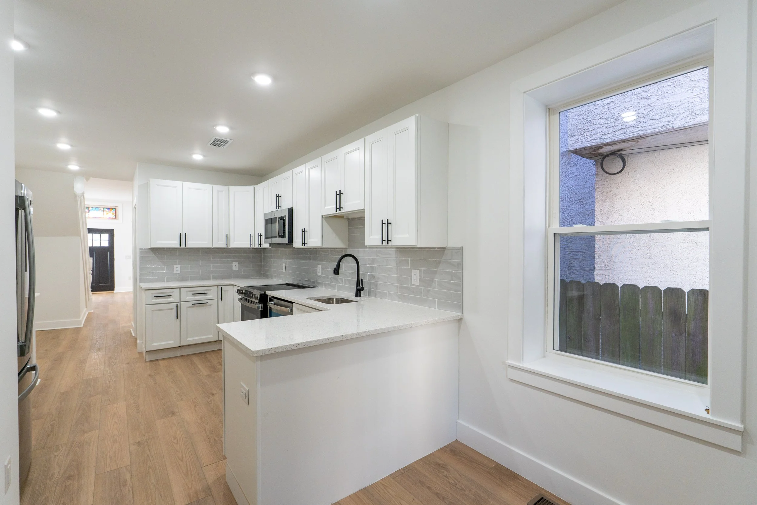 Modern kitchen with white cabinets, gray subway tile backsplash, stainless steel appliances, black faucet, and a large window overlooking a wooden fence outside.