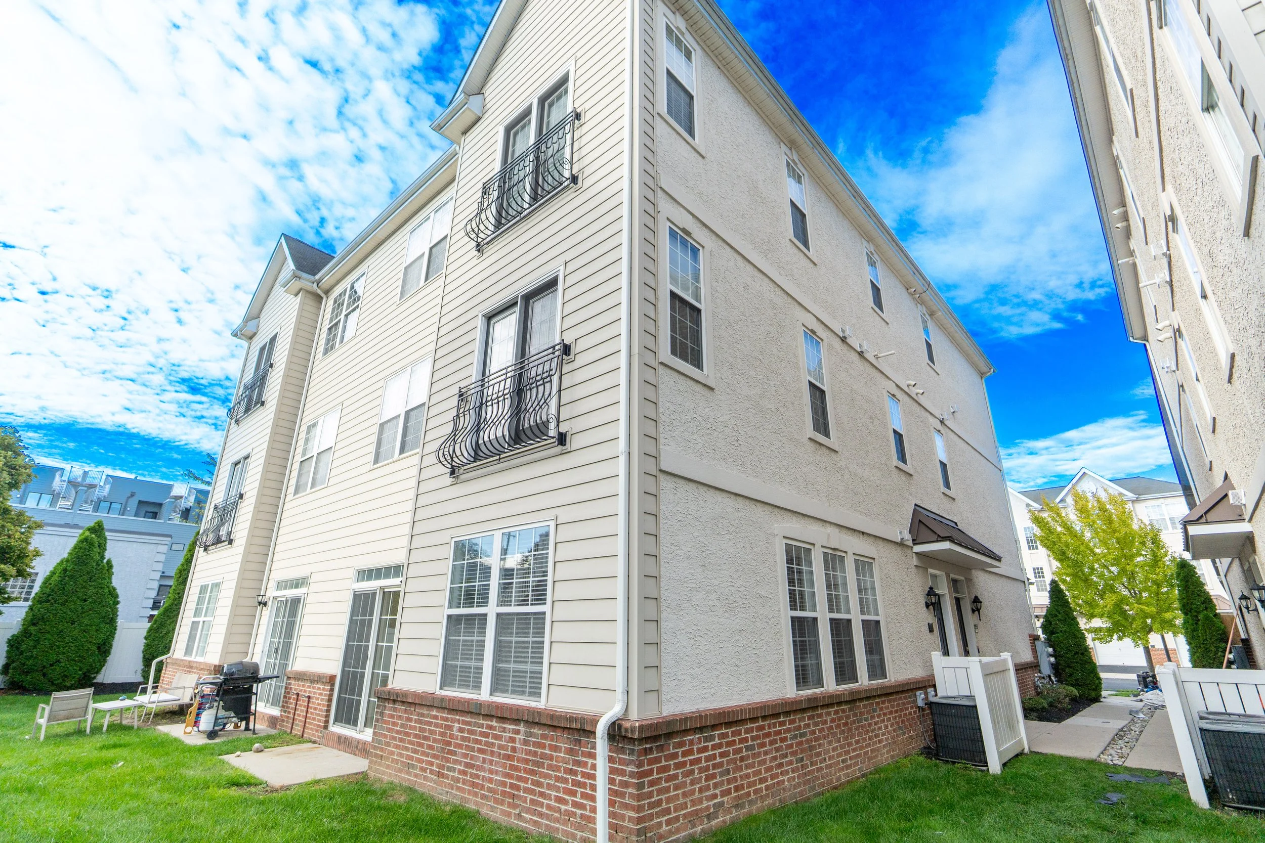 Exterior view of a multi-story residential building with cream siding, brick foundation, multiple windows, and small balcony railings under a partly cloudy blue sky.