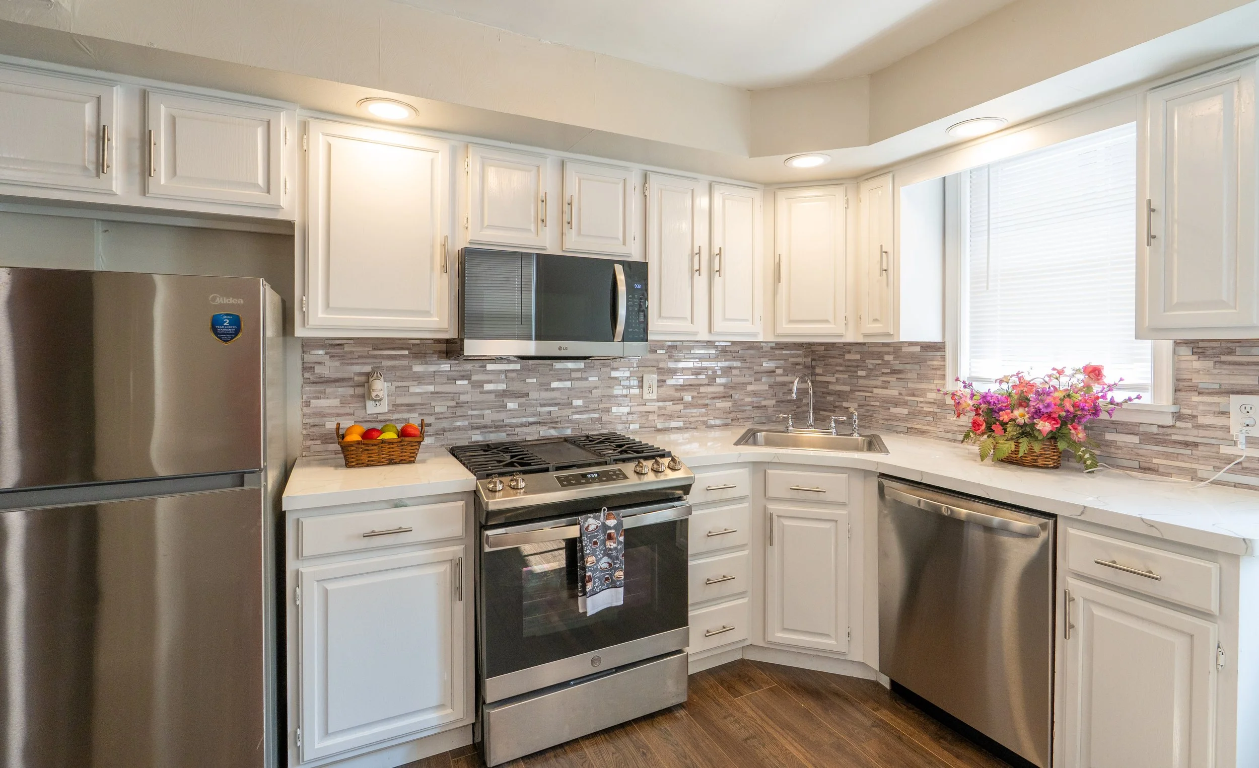 Kitchen with white cabinets, stainless steel refrigerator, microwave, dishwasher, and gas stove, beige tile backsplash, window with blinds, and a flower basket on the counter.