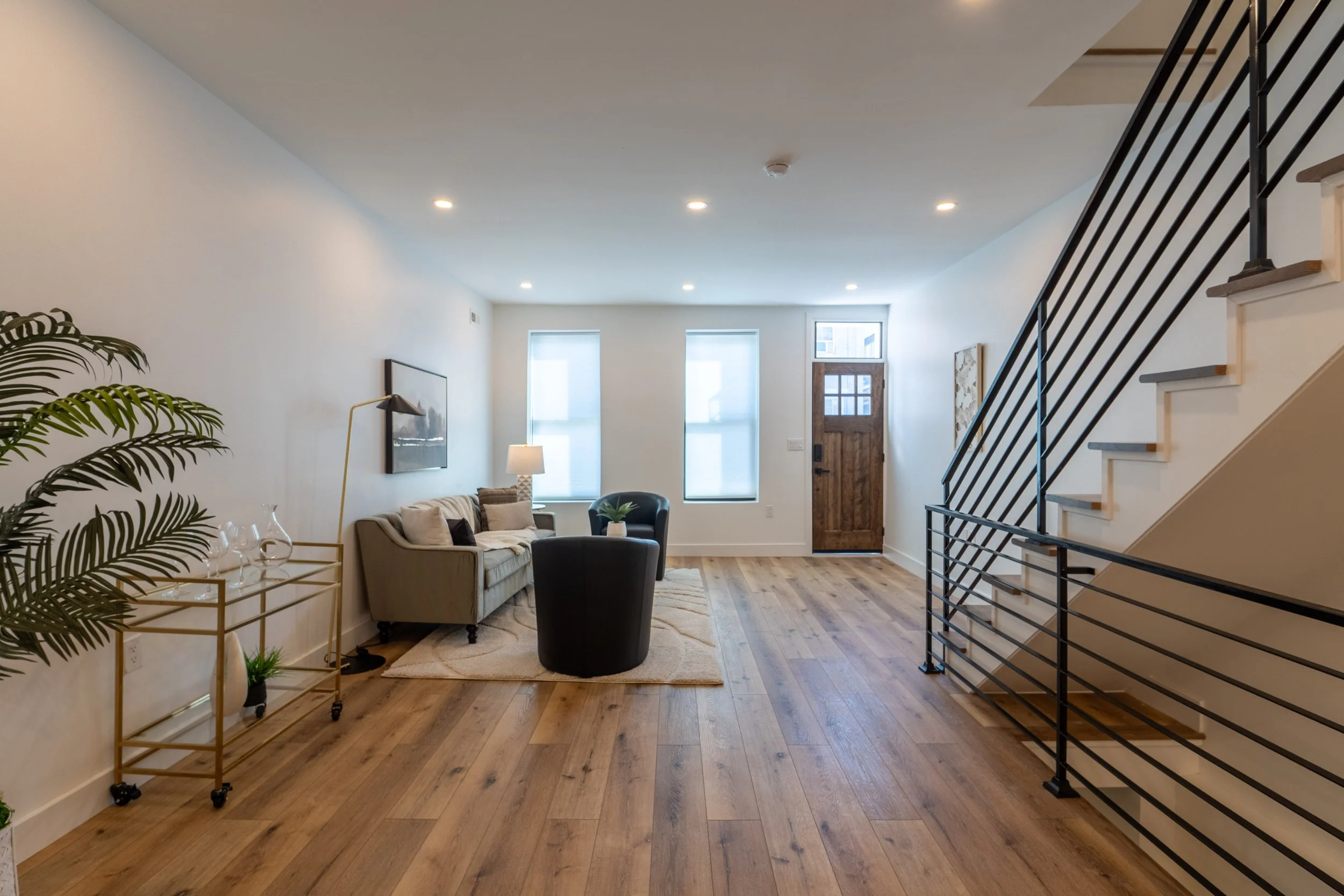 Living room with wooden floors, beige sofa with pillows, black armchair, lamps, artwork on white walls, staircase with black railing, front door, and windows with blinds.