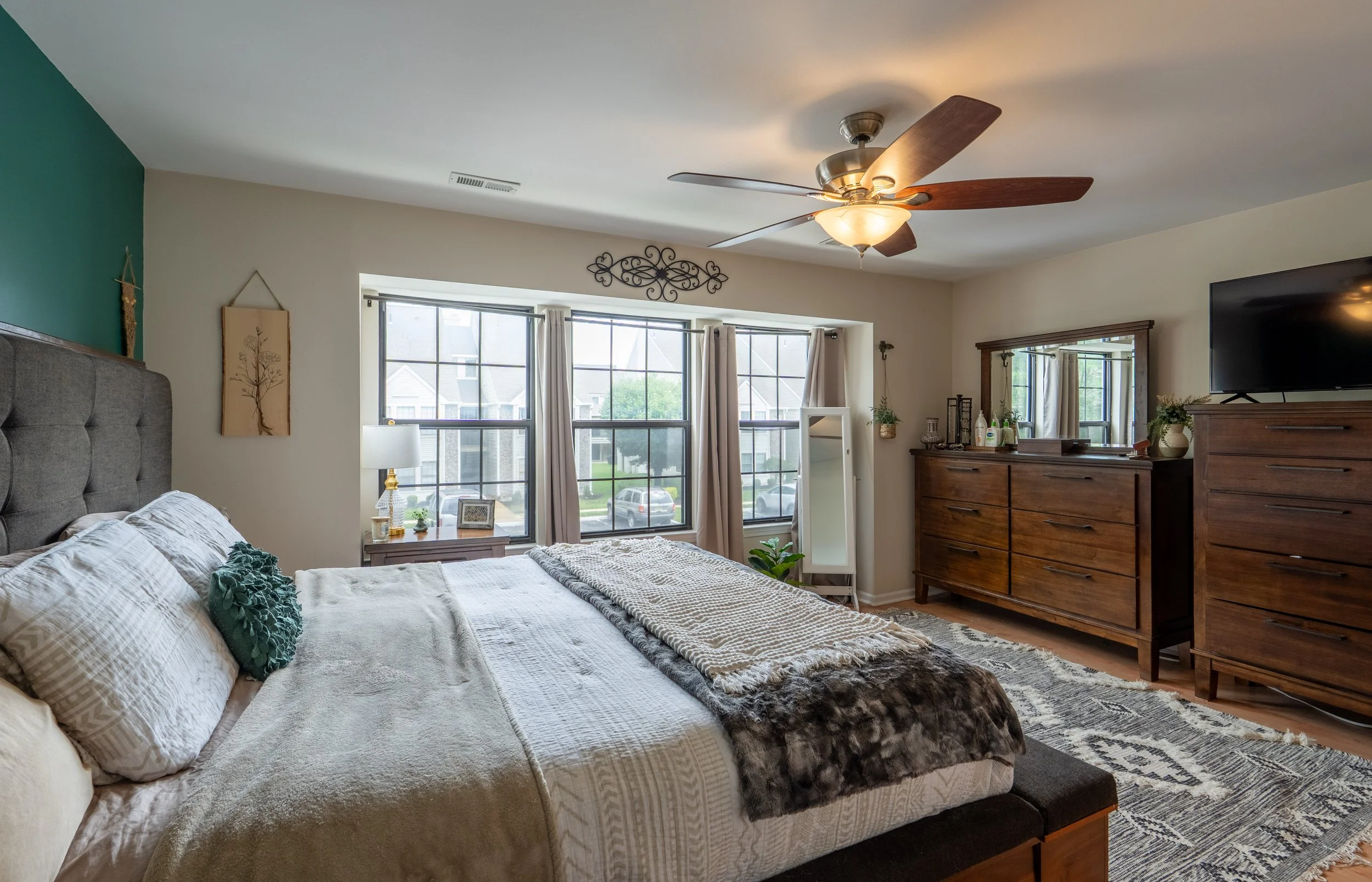 A bedroom with a large bed, wooden dresser, and a TV. The room has a ceiling fan, large windows with curtains, and various decorative items including plants and framed pictures.