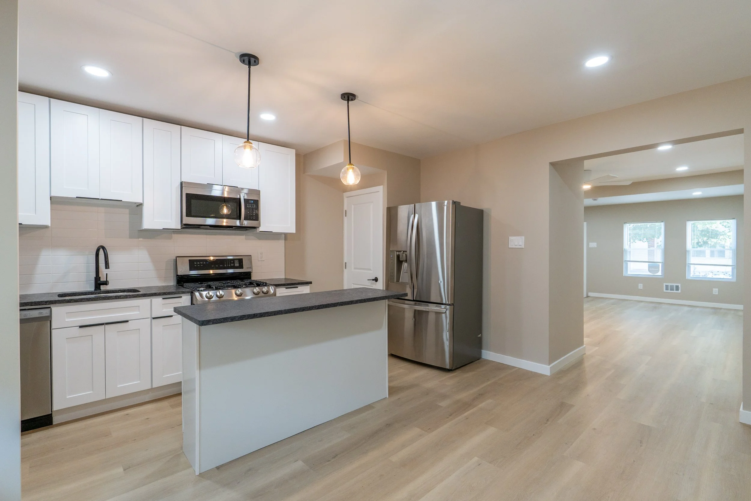 Modern kitchen with white cabinets, black countertops, stainless steel appliances including microwave, stove, and fridge, pendant lights, and adjacent living area with hardwood floors and windows.