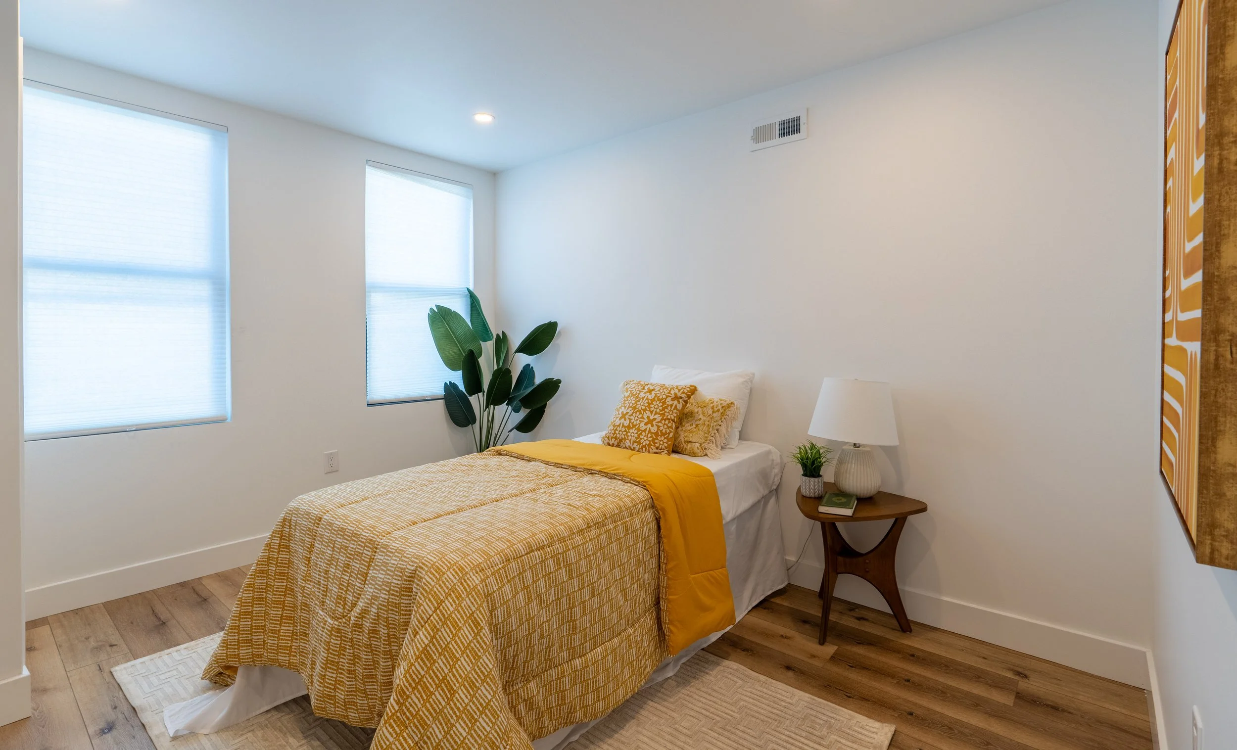 Bedroom with white walls, two windows with blinds, a bed with yellow bedding and pillows, a wooden nightstand with a lamp and a potted plant, a large leafy plant near the window, hardwood floor, and a wall art piece.