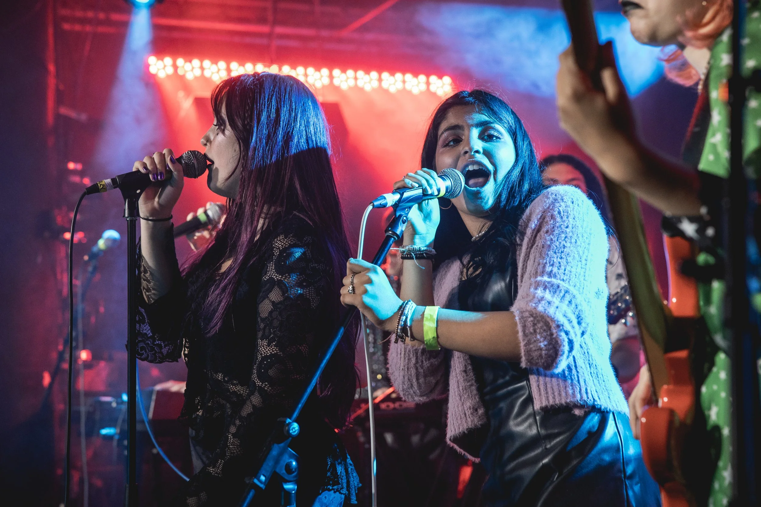Two young women singing into microphones on a stage with colorful lights, one with long dark hair and lace top, the other with shoulder-length black hair and a pink sweater.