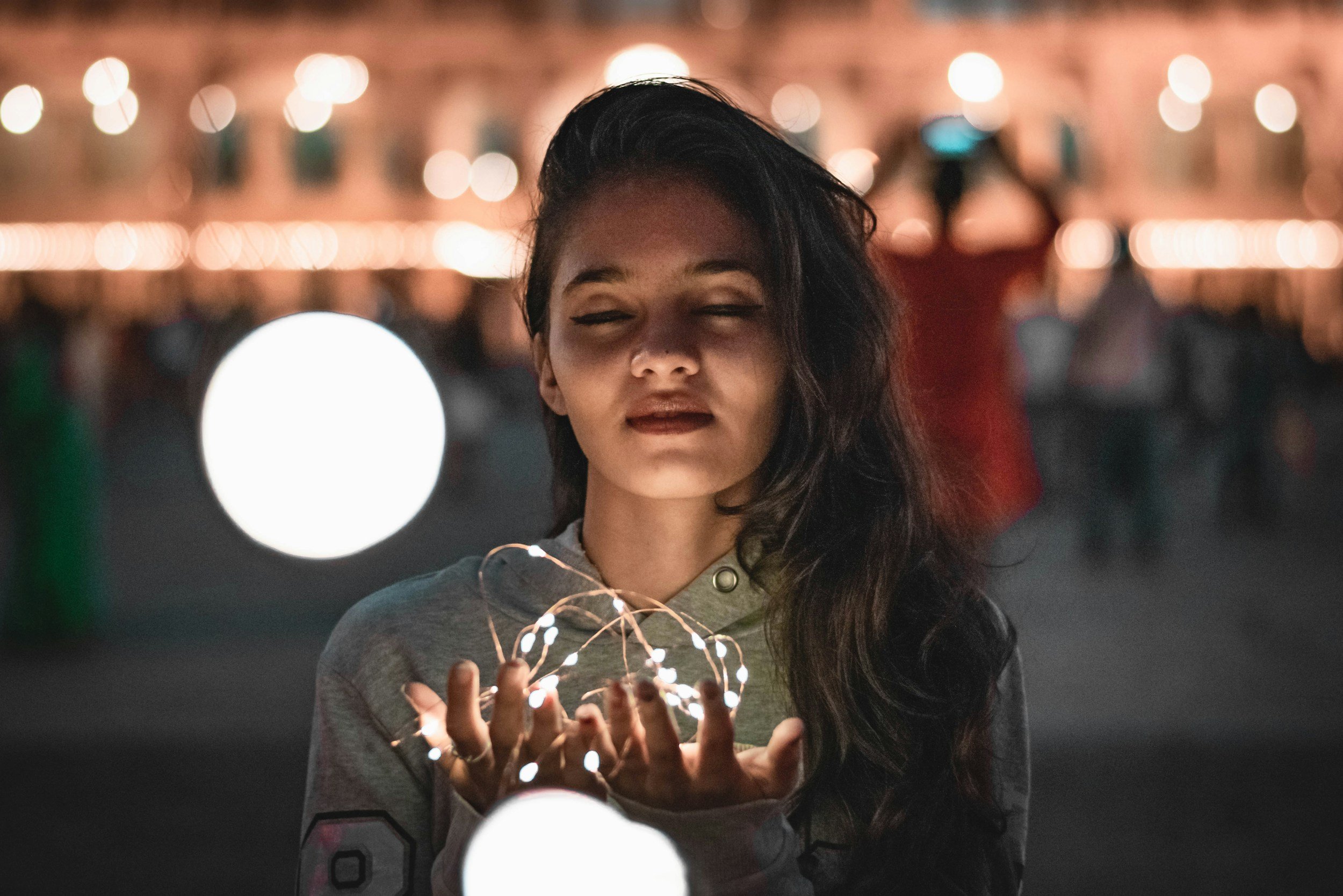 A young woman with long dark hair, eyes closed, holding string lights in her hands at night, illuminated in a festive or outdoor setting with blurred background lights.