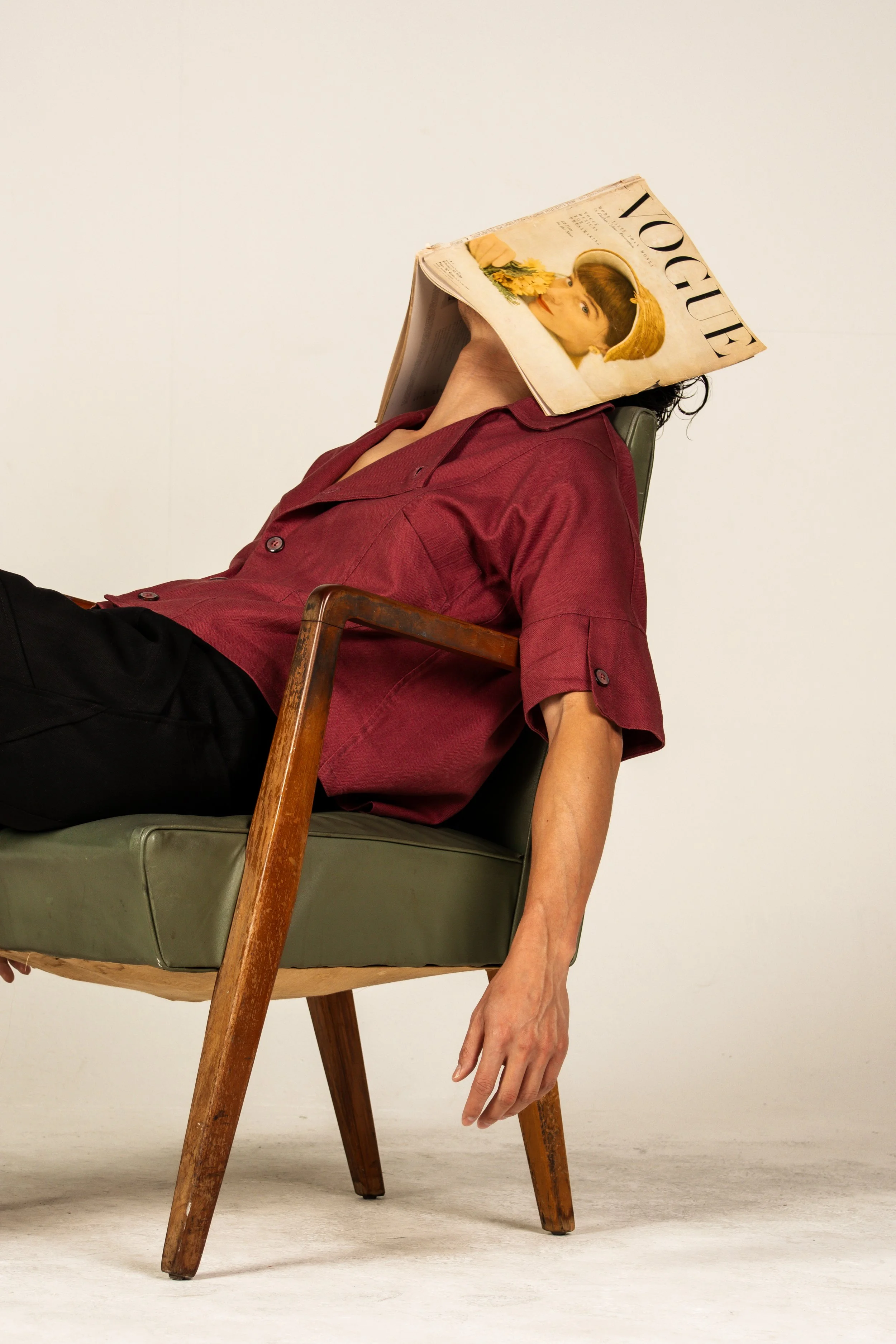 A person is reclining in a green armchair with a Vogue magazine covering their face. The person is wearing a red shirt and dark pants.