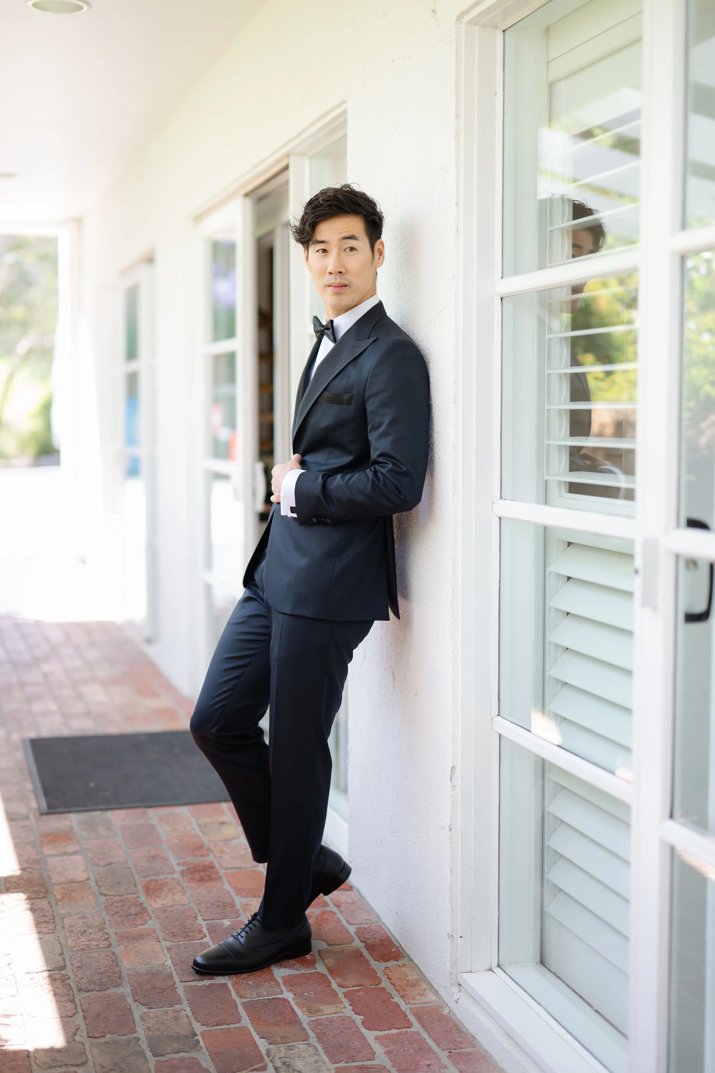 Ray Yee in a navy tuxedo leaning against a white wall outside