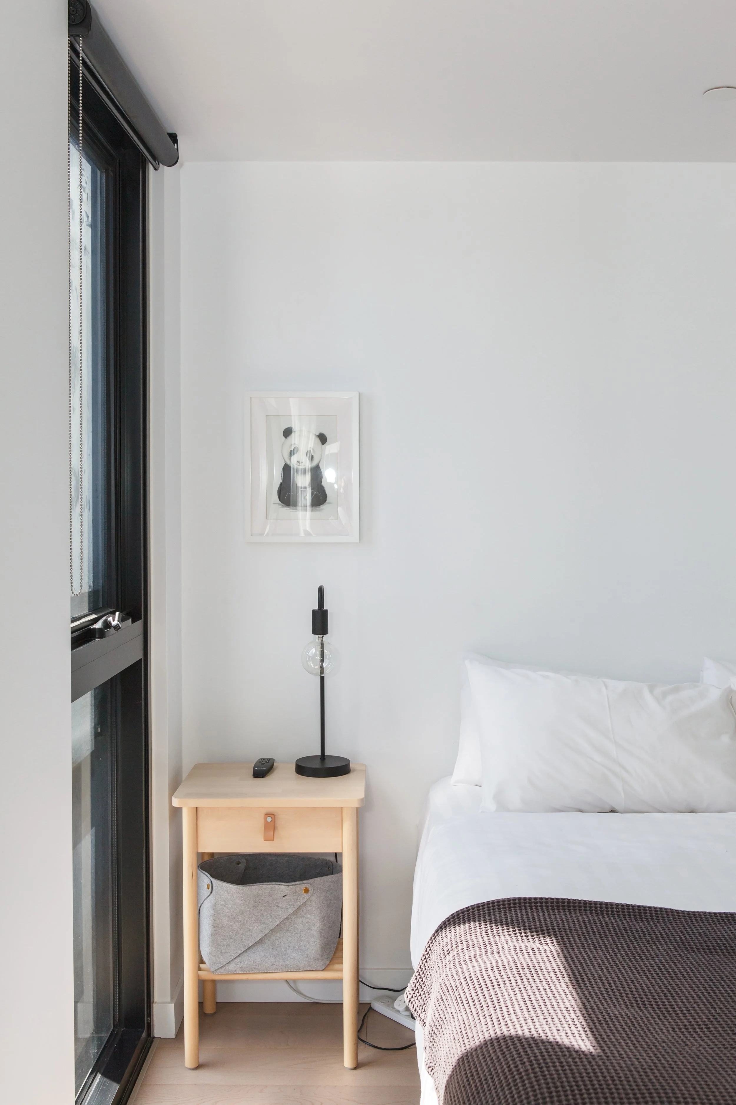 A bedroom corner with a bed dressed in white bedding, a small light wooden nightstand, a black table lamp, a TV remote, a gray fabric basket inside the nightstand shelf, a framed panda picture on the wall, and a tall window with black trim and vertical blinds.