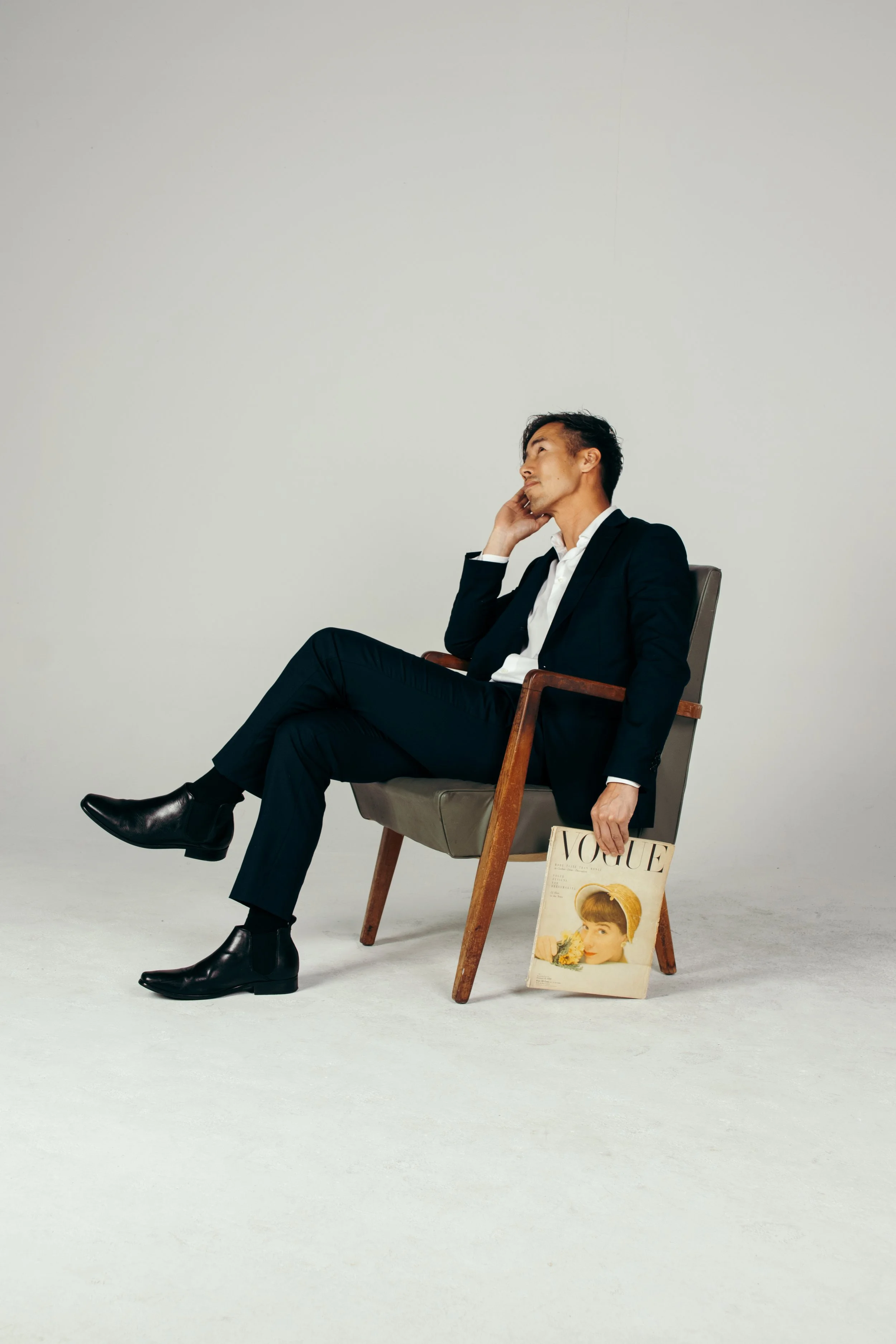 A man in a black suit and white shirt sitting on a wooden armchair, resting his head on his hand, with a Vogue magazine placed on the floor beside him.