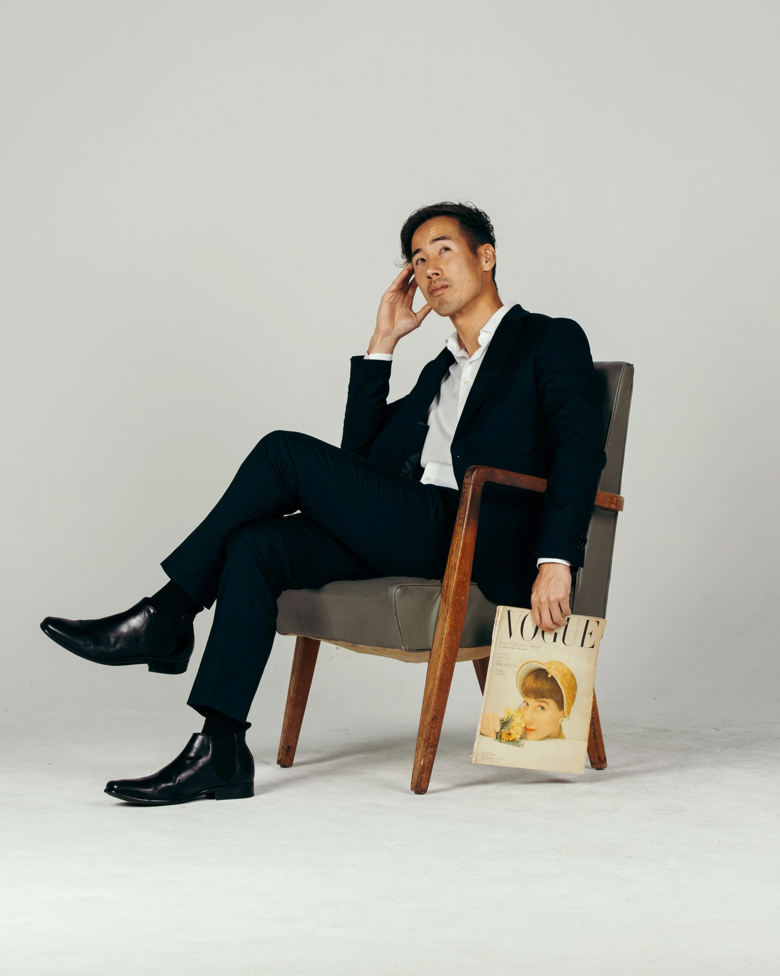 A man in a black suit sitting on a wooden chair in a studio with a Vogue magazine hanging from the armrest. He's holding his head with his left hand and looking slightly puzzled.