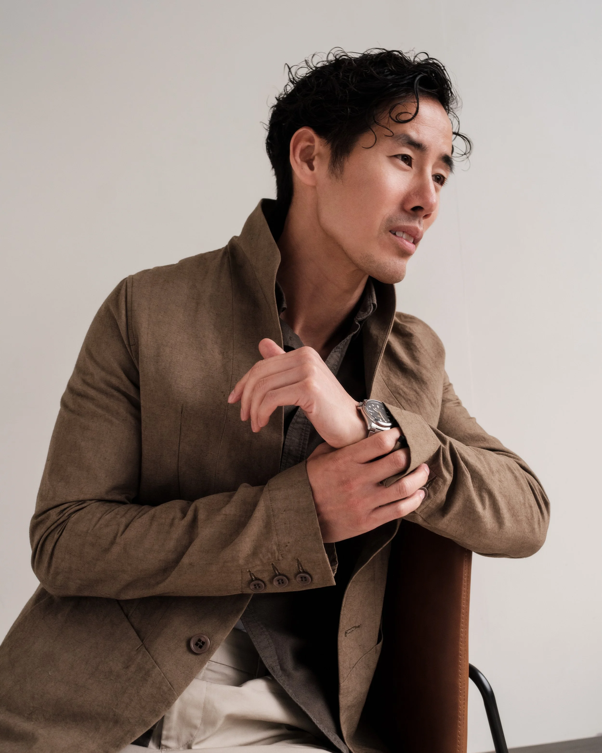 A man with dark, curly hair wearing a brown jacket, sitting in a chair, with his arms crossed, against a plain white wall.