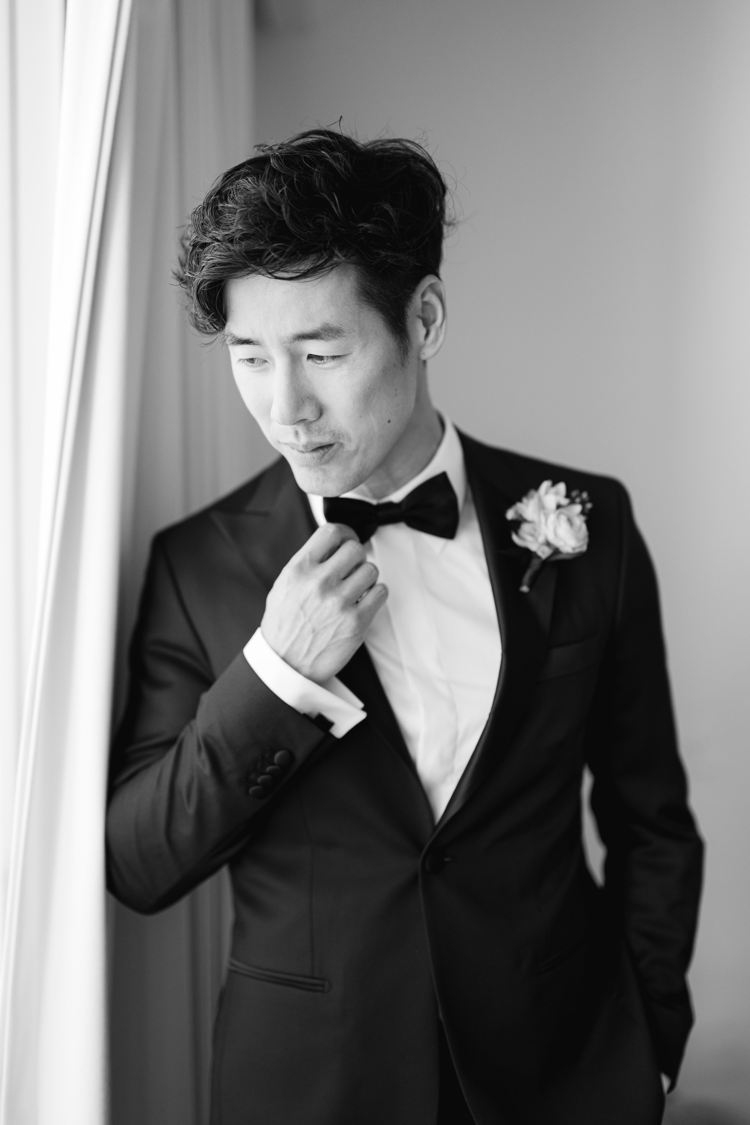Ray Yee in a tuxedo adjusting his bow tie by a window, black and white