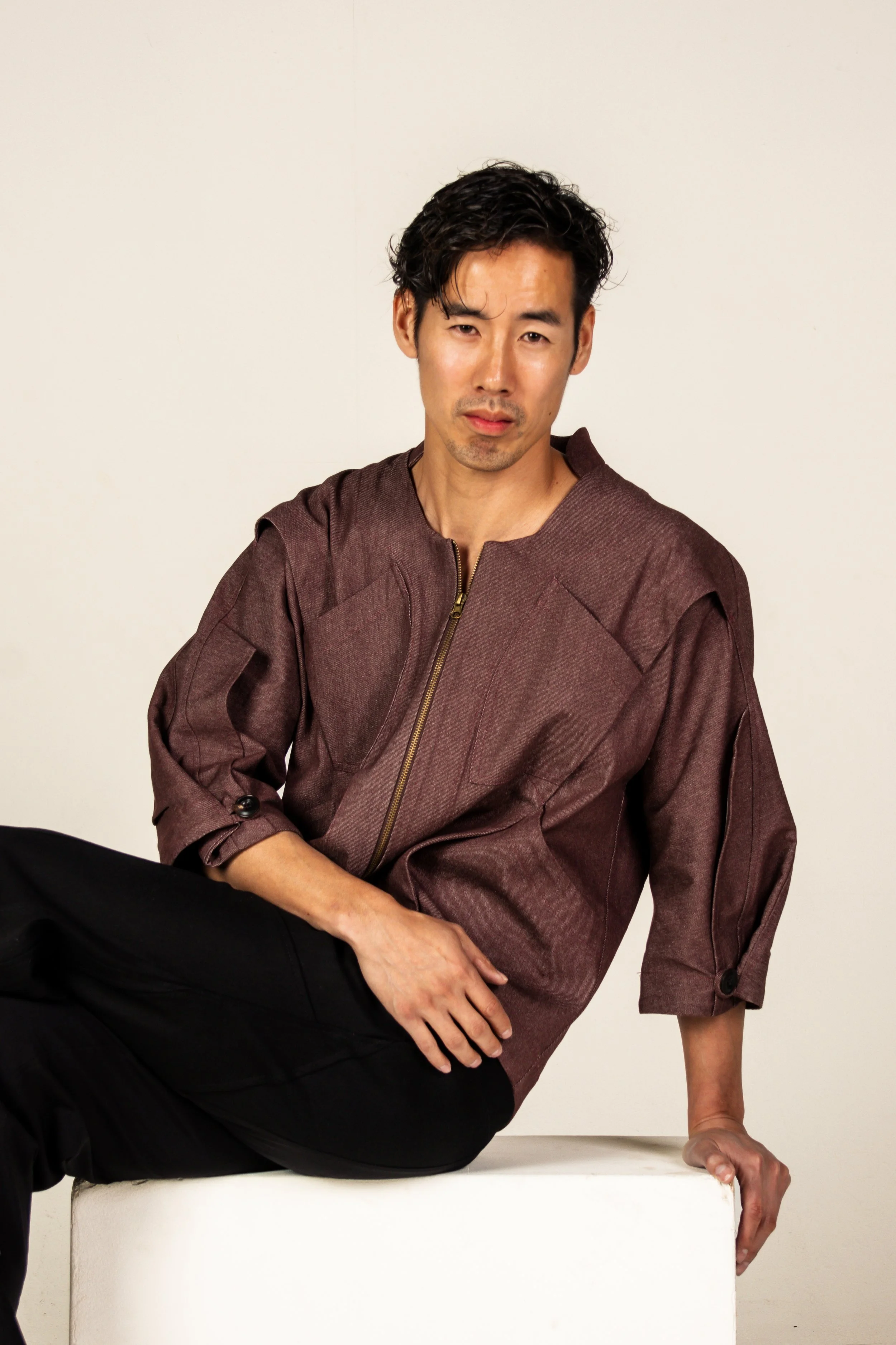 A man with black hair, wearing a brown zip-up jacket and black pants, is sitting on a white platform against a plain white background, looking at the camera with a neutral expression.