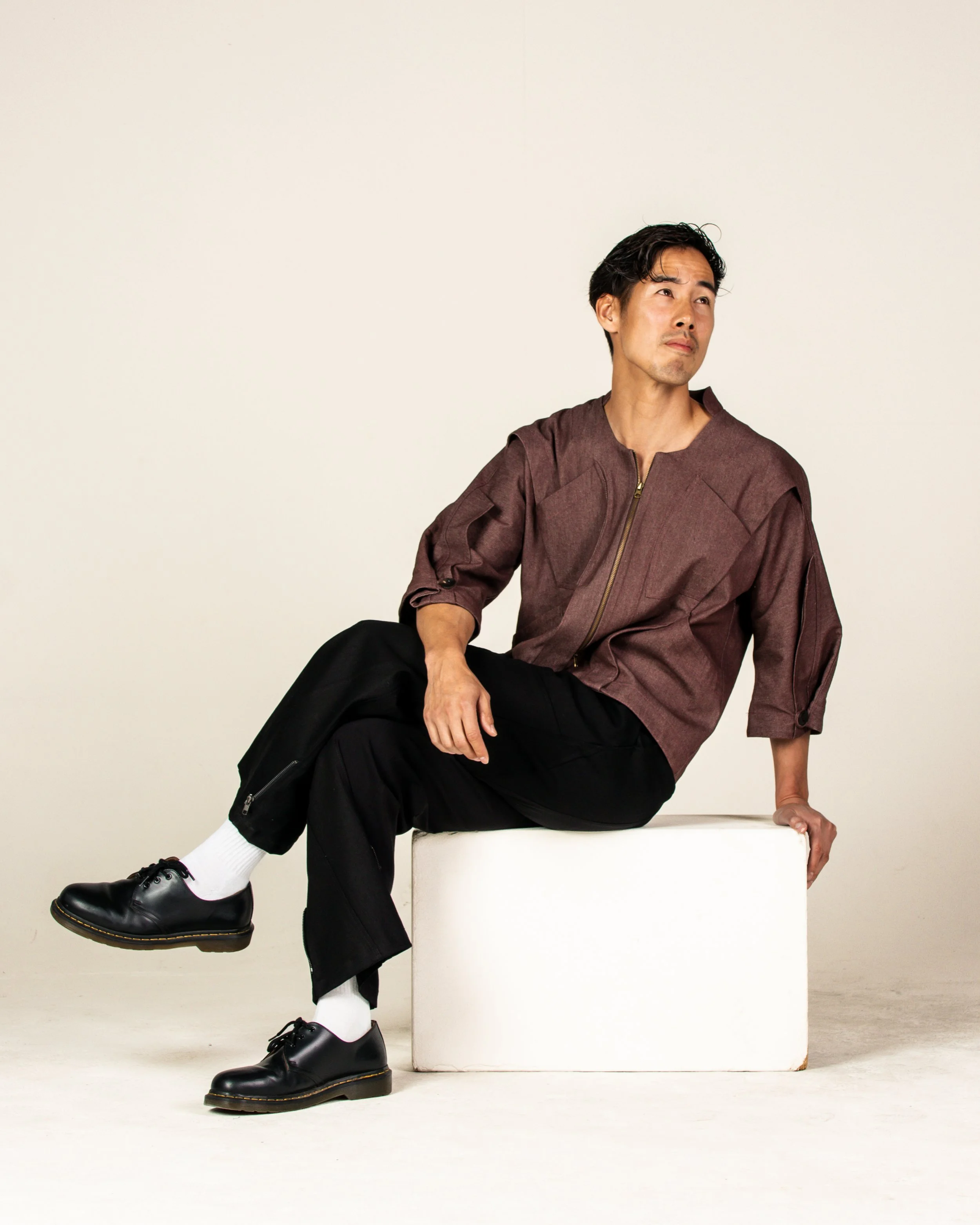 A man sitting on a white cube, wearing a brown jacket, black pants, white socks, and black shoes, against a plain off-white background.