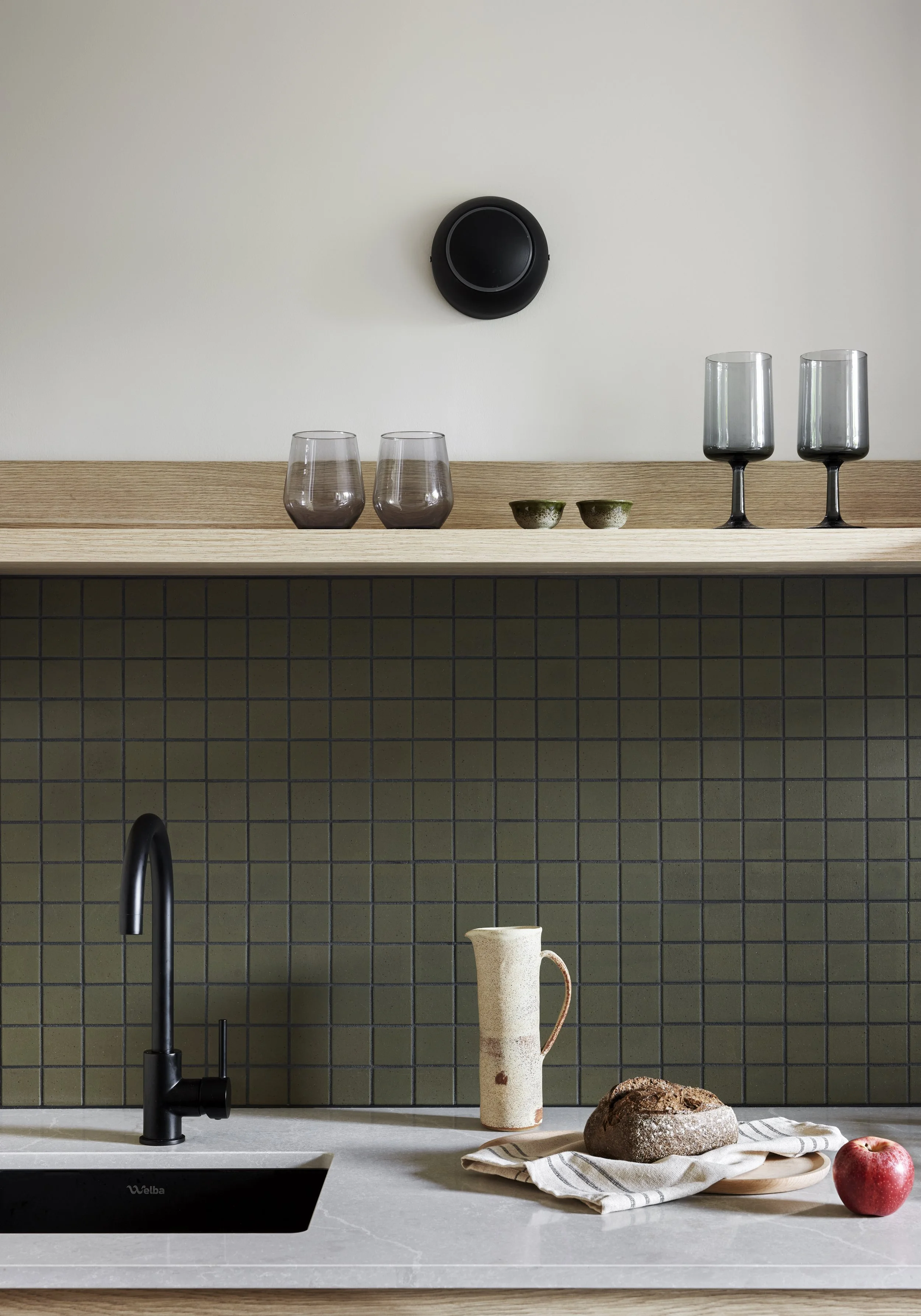 Modern kitchen with black faucet, beige pitcher, loaf of bread, apple on a white towel, green tiled backsplash, and glassware on wooden shelf above.