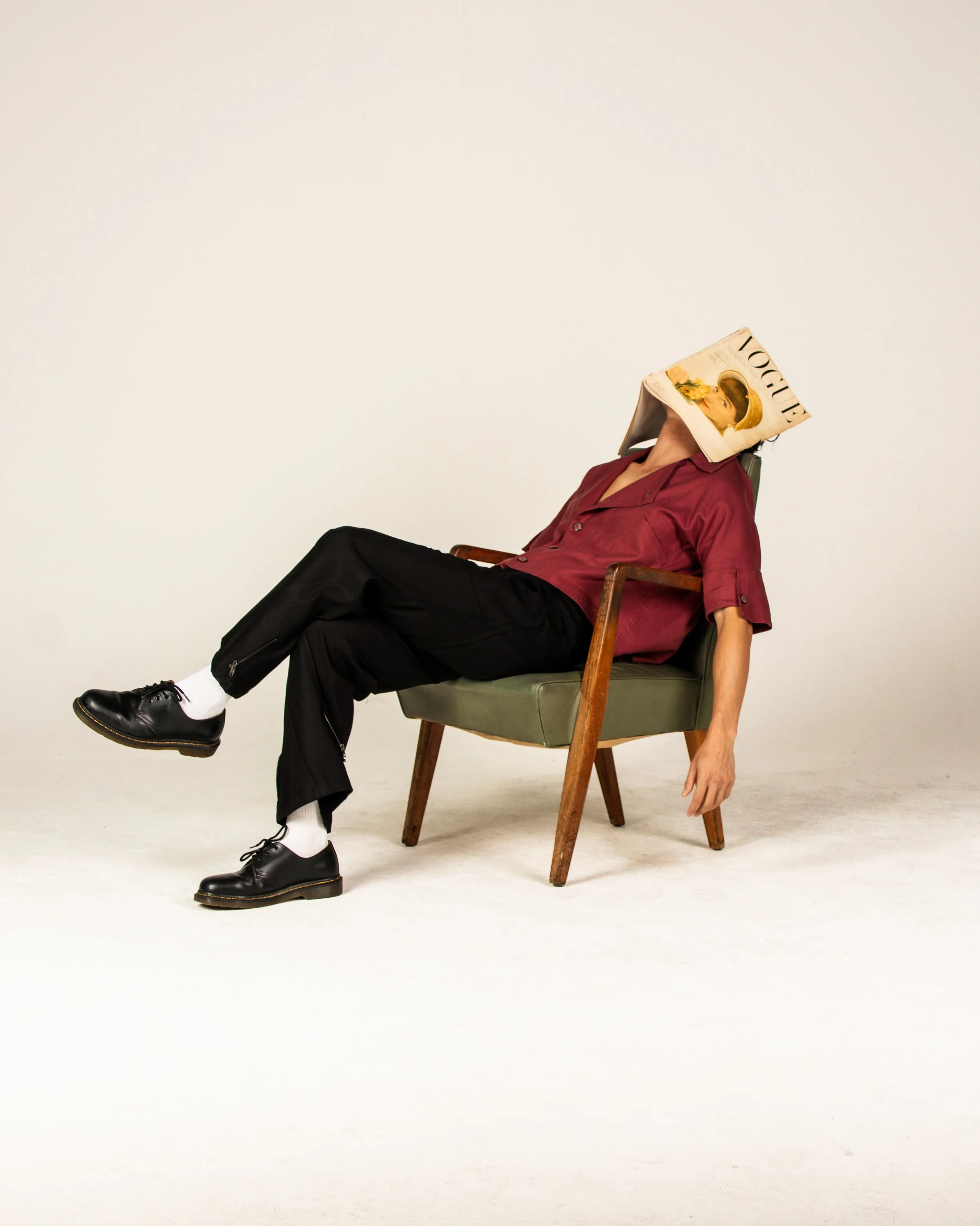 Person sitting in a green chair with a Vogue magazine covering their face, wearing a red shirt, black pants, white socks, and black shoes, with a plain white background.