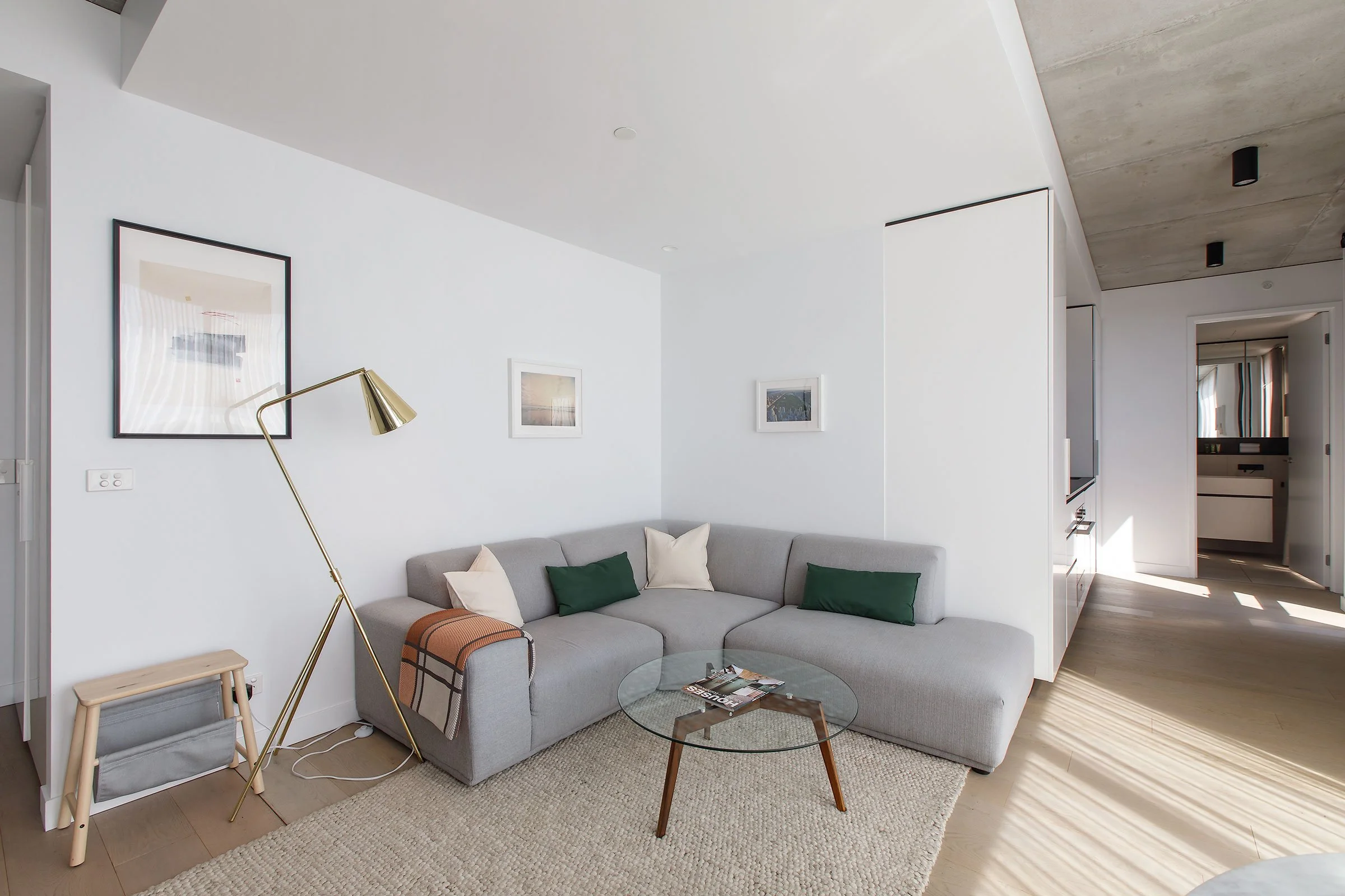 Modern living room with a light gray sectional sofa, white and green pillows, a round glass coffee table, a beige rug, and wall art, with sunlight streaming in.