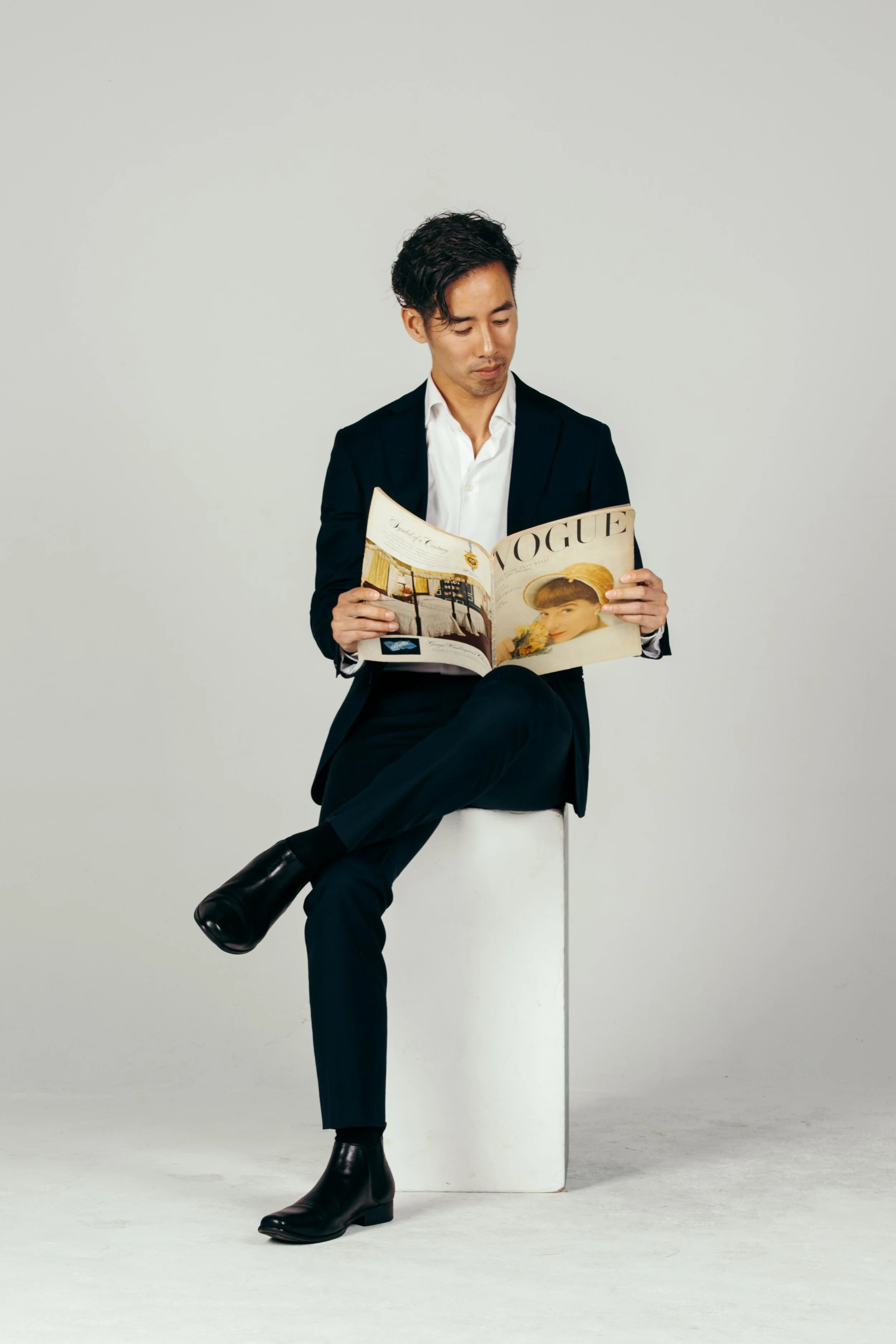A man in a black suit with a white shirt sitting on a white cube, reading a Vogue magazine.