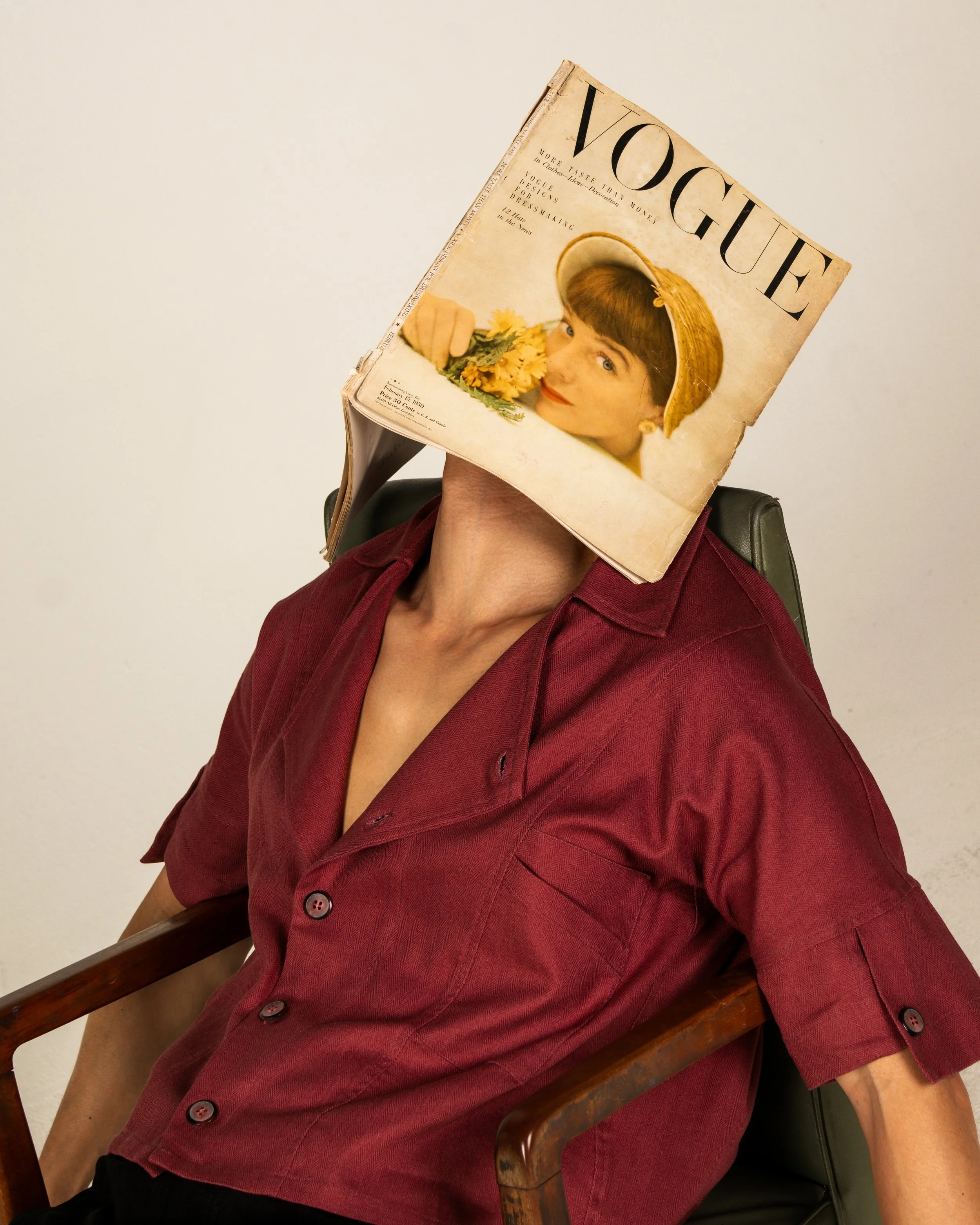 Person sitting in a chair with a vintage Vogue magazine cover as a mask. The magazine cover features a woman with a yellow hat and flowers.