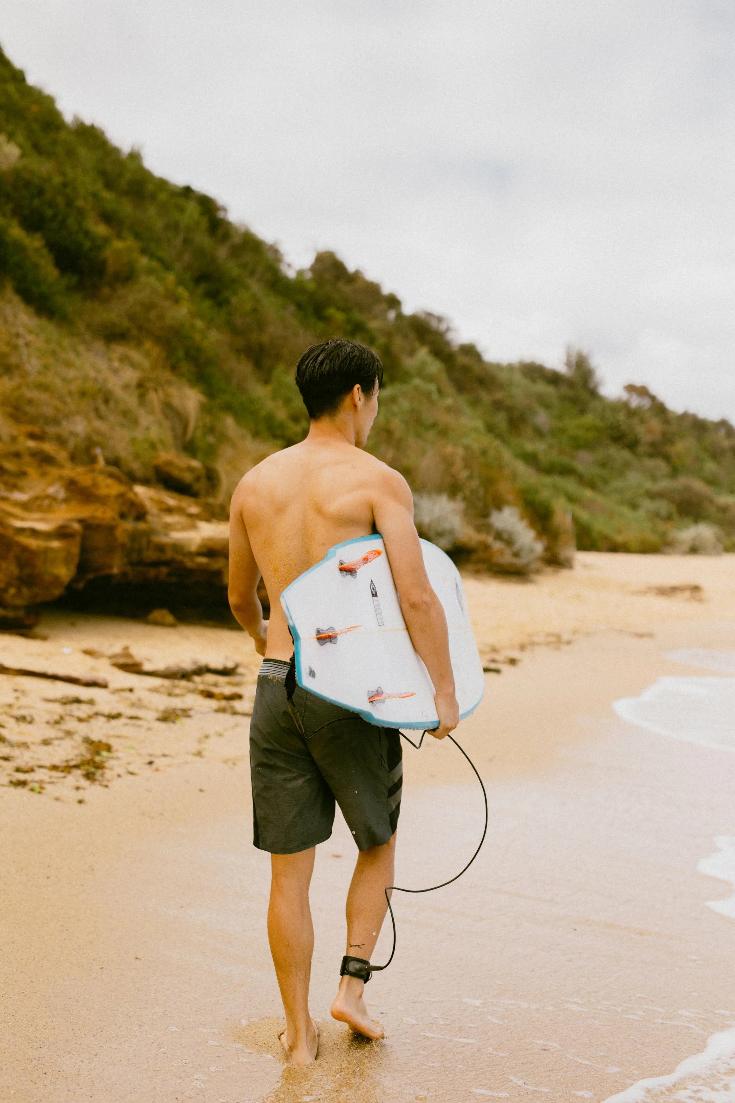 Ray Yee, Asian male model in Melbourne and surf coach, carrying a surfboard on the beach