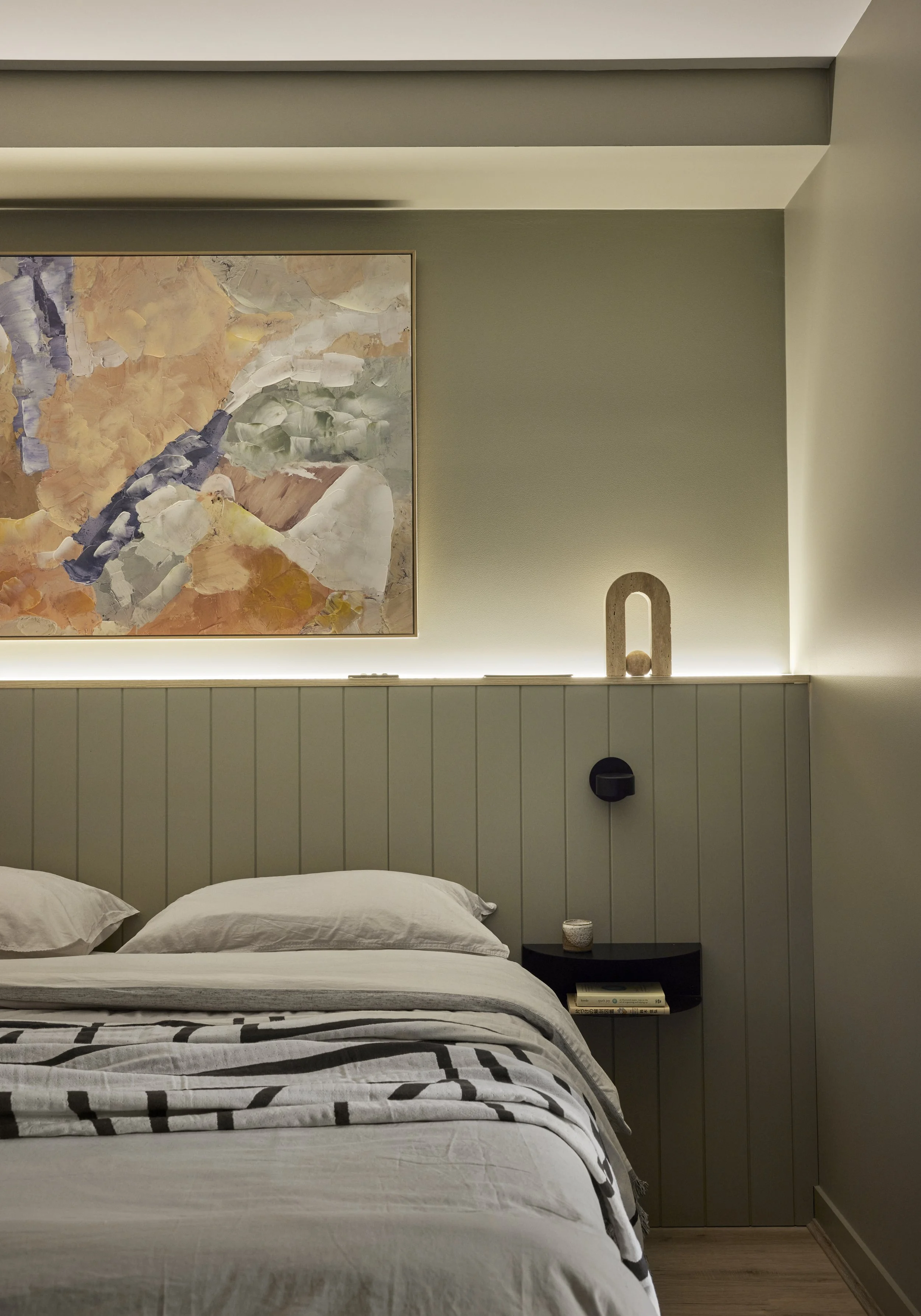 A bedroom with a bed that has white sheets and pillows, a black bedside table with a small decor item and books, a large abstract painting on the wall, and subtle lighting behind the headboard and on the wall.