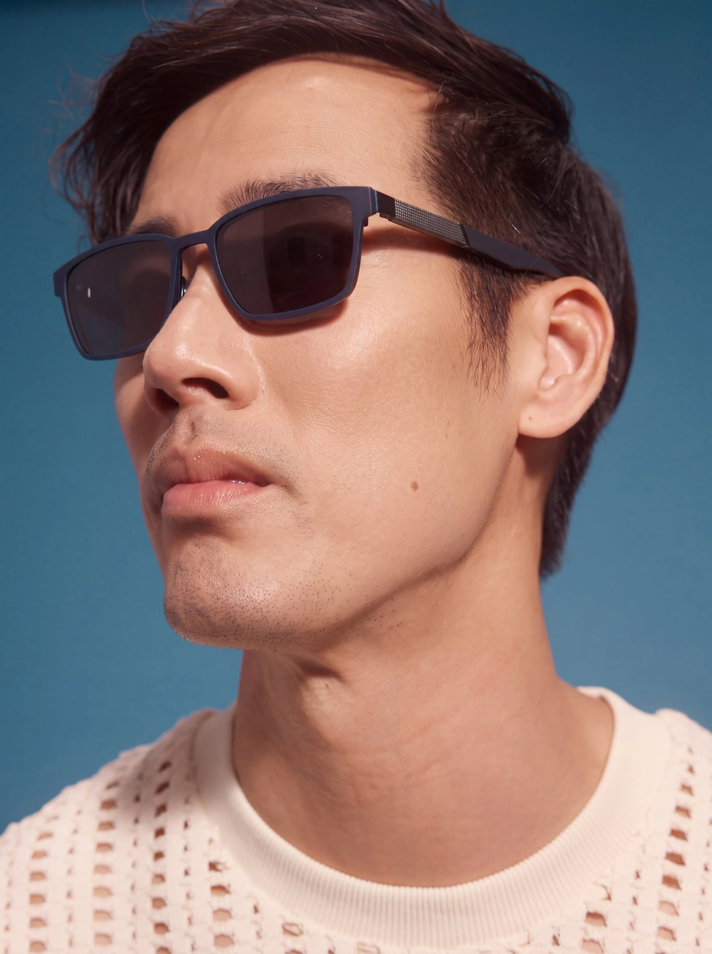 Ray Yee wearing dark sunglasses and a white knit top against a blue background, Convertibles eyewear campaign