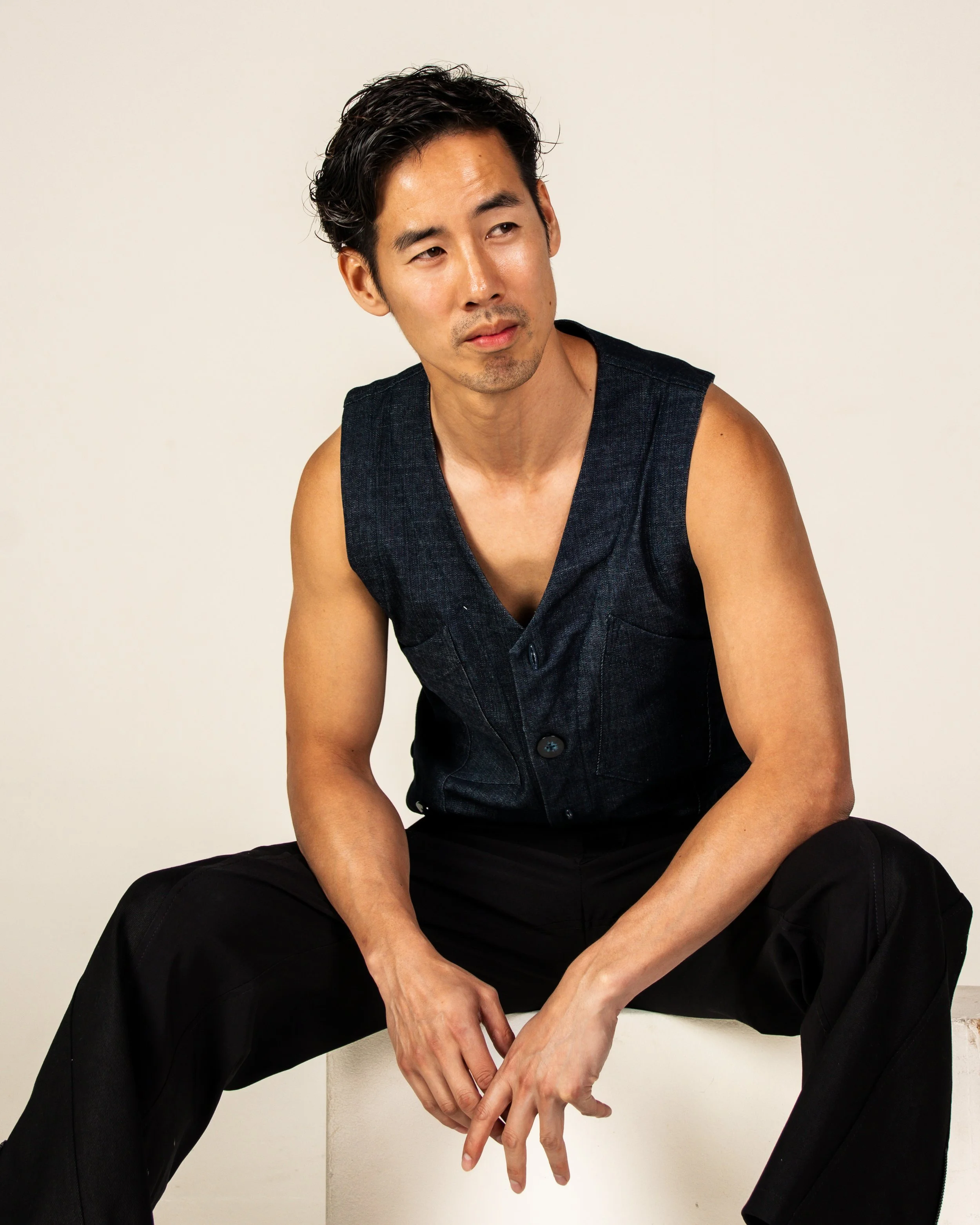 A man sitting on a white block, wearing a dark sleeveless shirt and black pants, looking slightly to the side with a neutral expression.