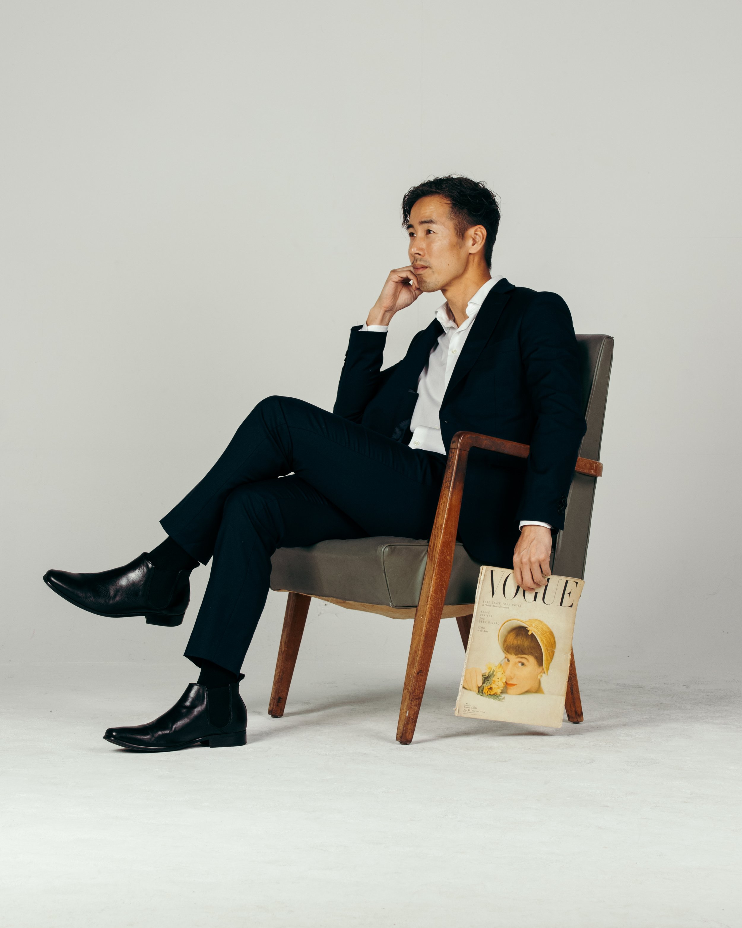A man in a black suit sitting in a chair, holding a Vogue magazine, in a minimalistic setting.