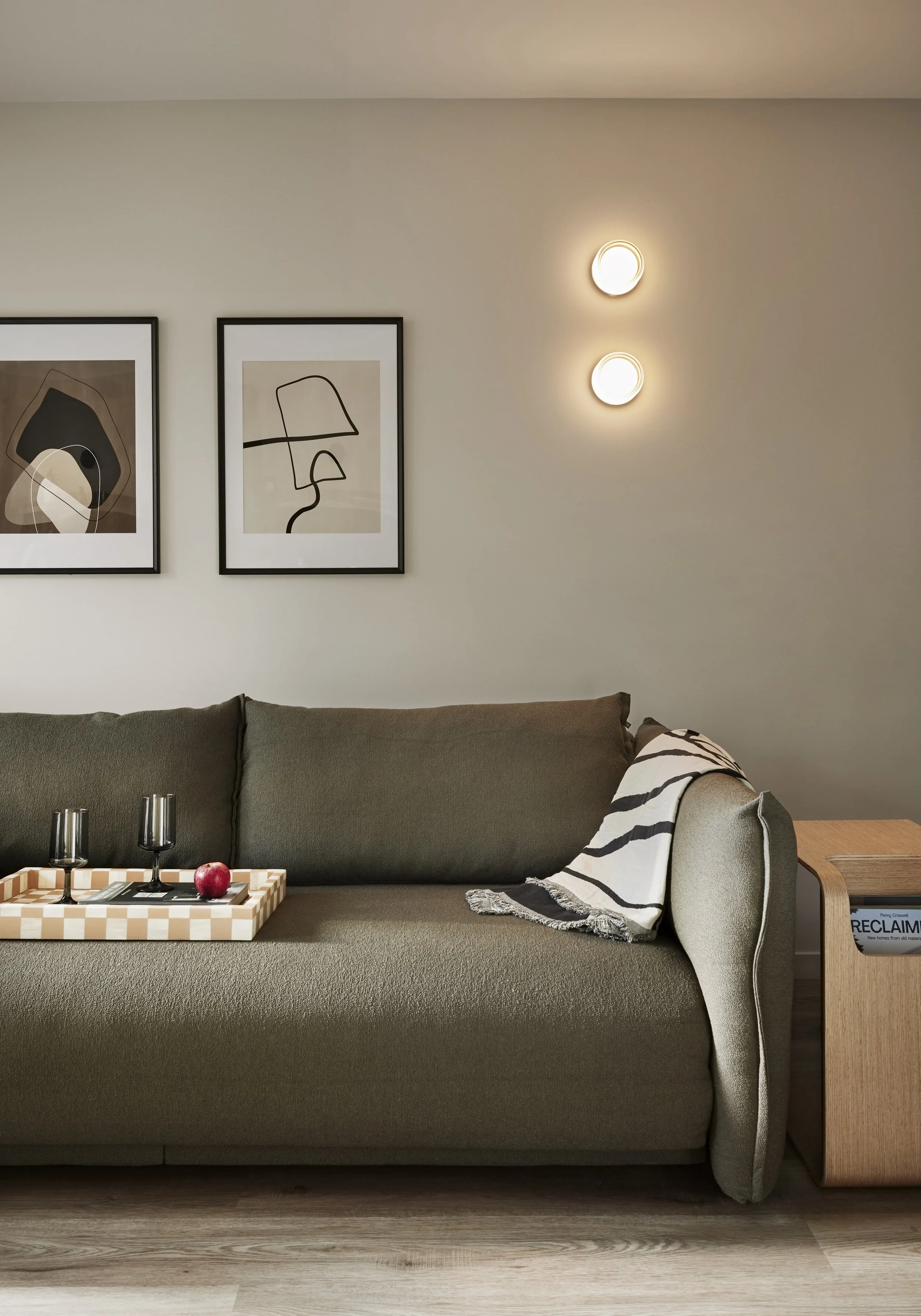 Living room with a green sofa, black and beige abstract artwork, a blanket, and a wooden side table with a magazine. Two round wall lights are on the wall.