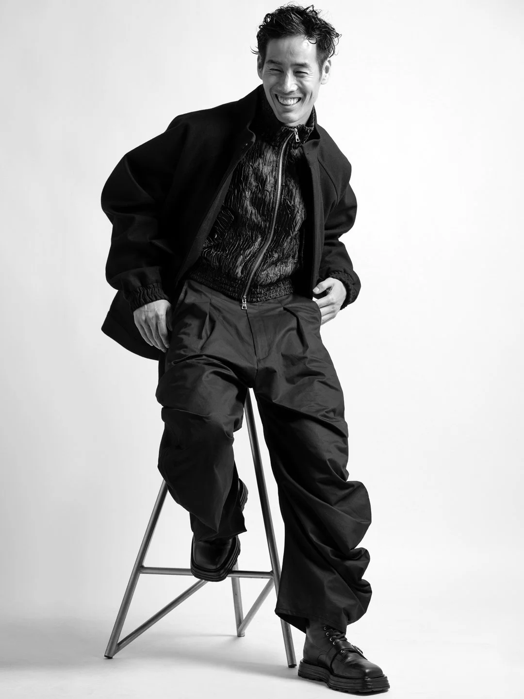 Ray Yee laughing, seated on a stool in a bomber jacket and wide trousers, black and white