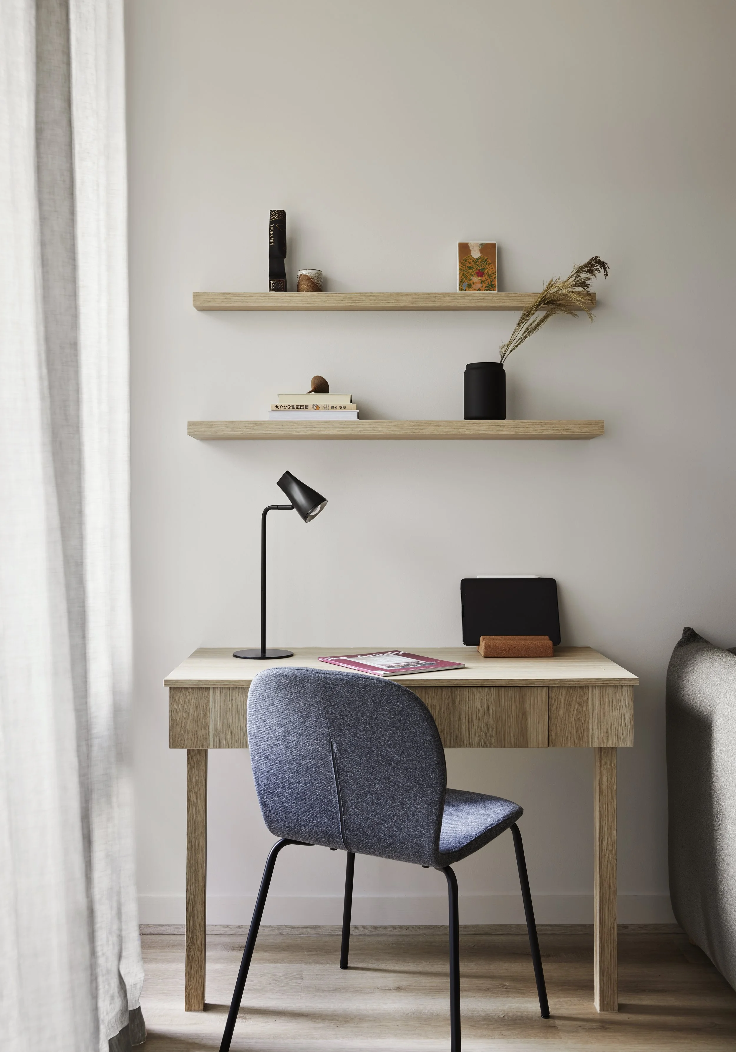 A minimalist home workspace with a light wood desk, gray upholstered chair, black desk lamp, and floating shelves decorated with books, a small painting, and a black vase with dried grasses, near a window with white curtains.