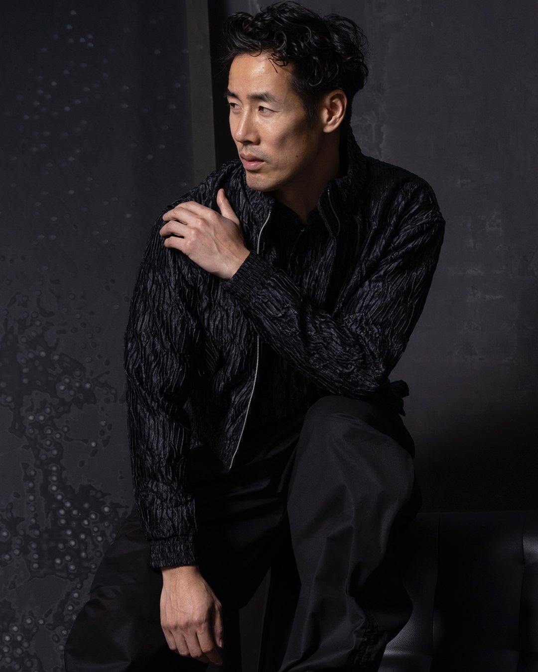 Ray Yee in a black textured jacket and black trousers against a dark wall