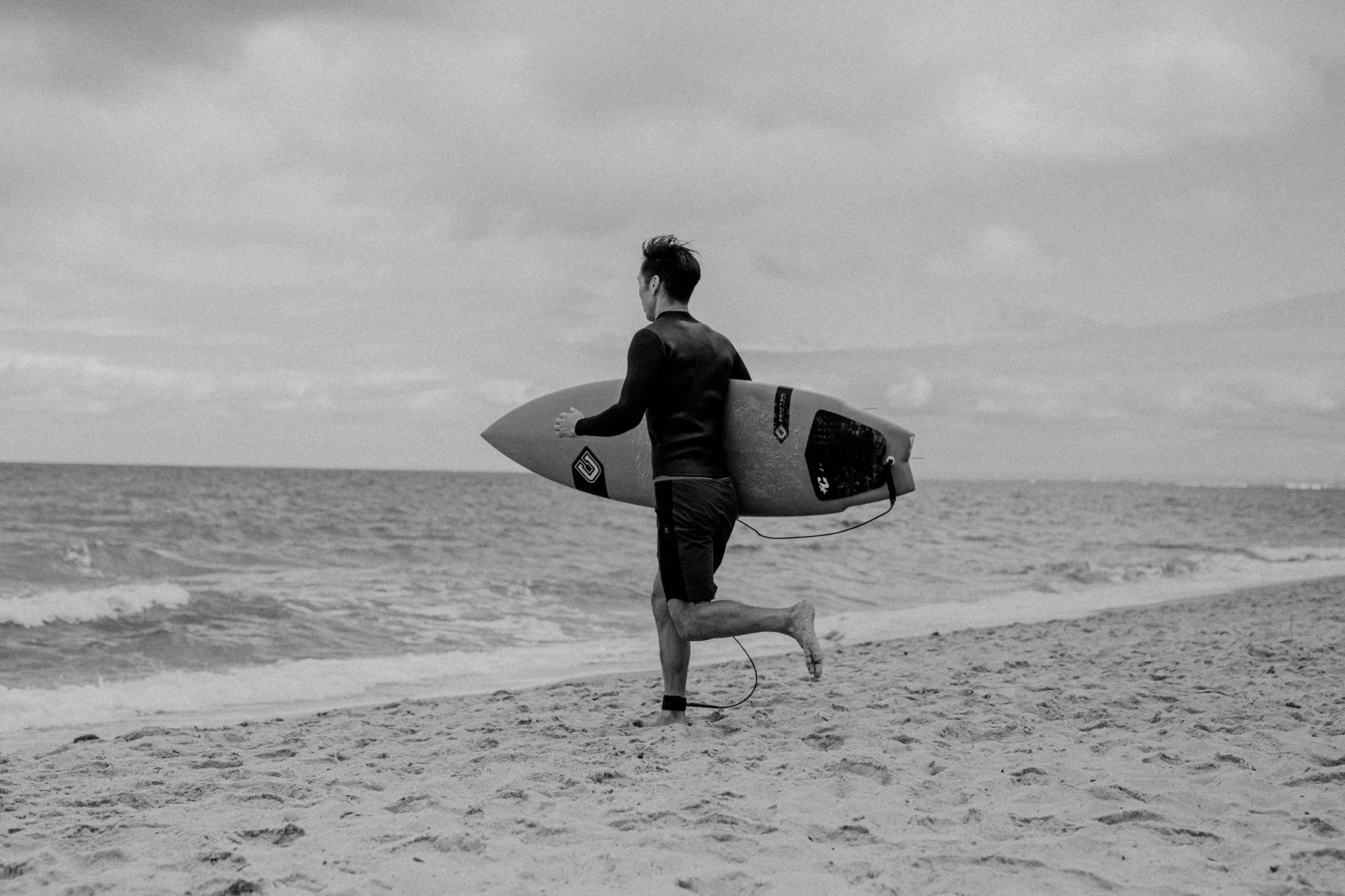 A man running on the beach holding a surfboard under his arm, with ocean waves in the background, in black and white.