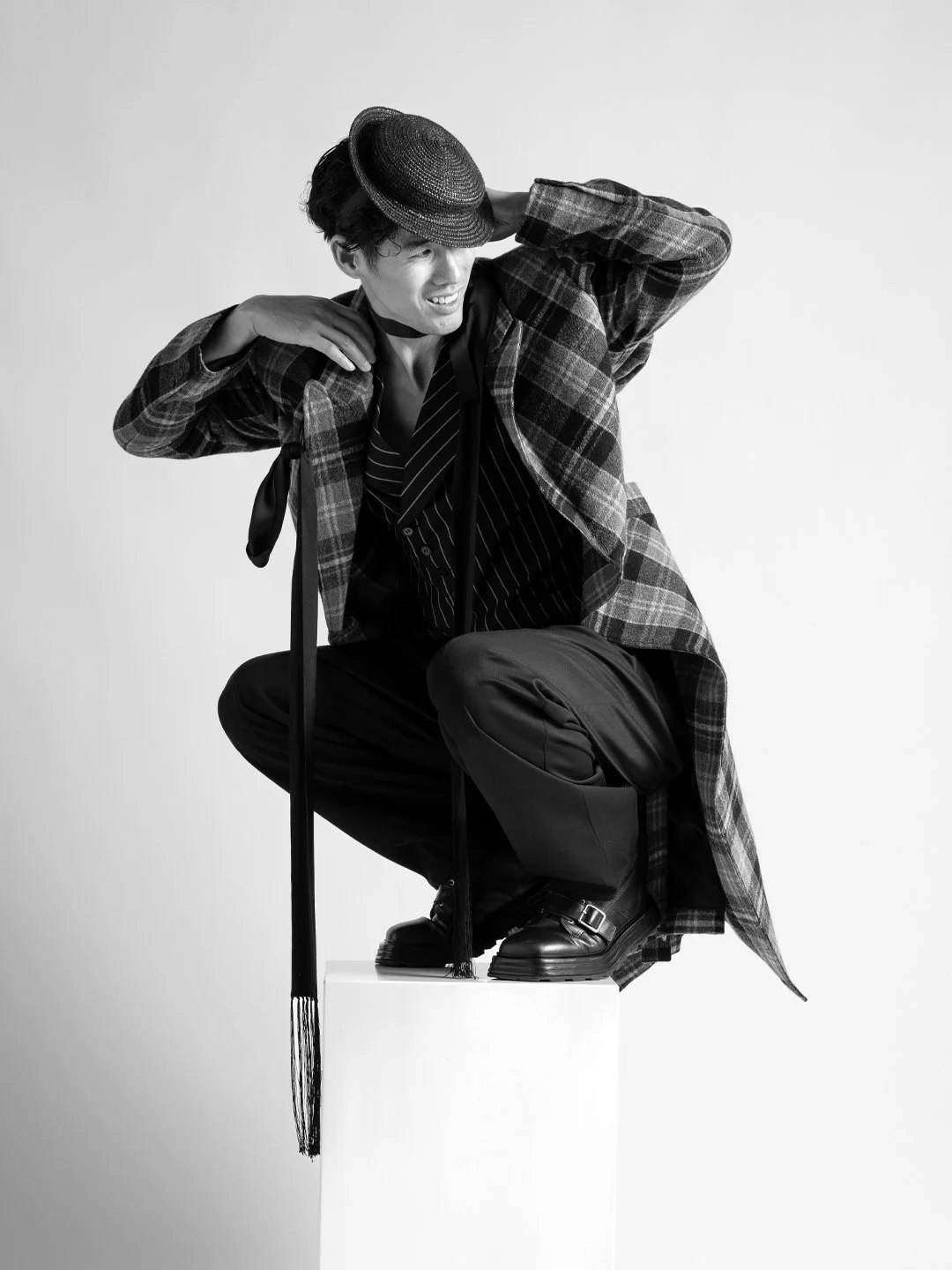 Ray Yee in a plaid coat and hat, crouching on a white block
