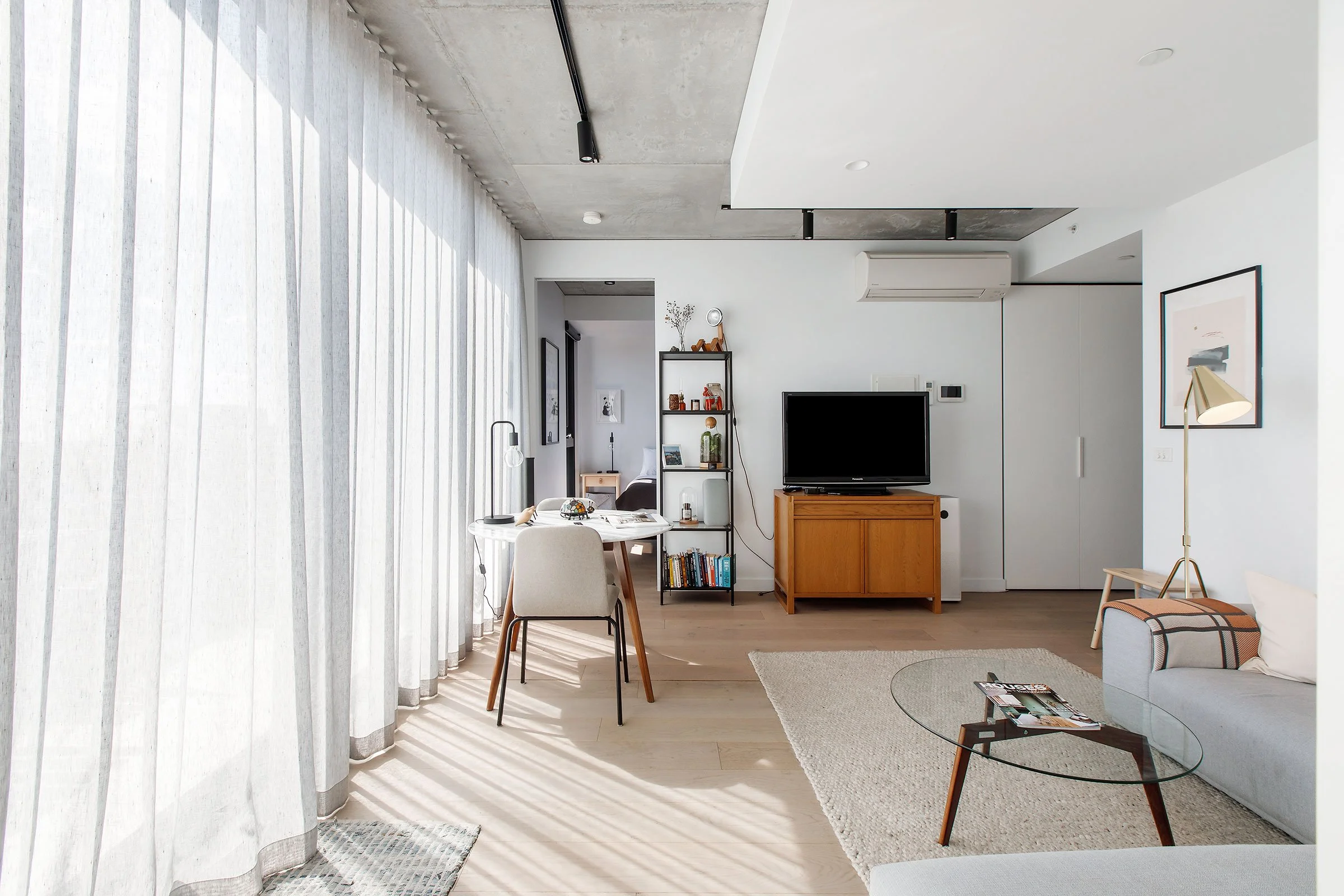 Modern living room with white walls, light wood flooring, sheer curtains, a small dining table with a chair, a TV on a wooden stand, a bookcase, a sofa, and a glass coffee table.