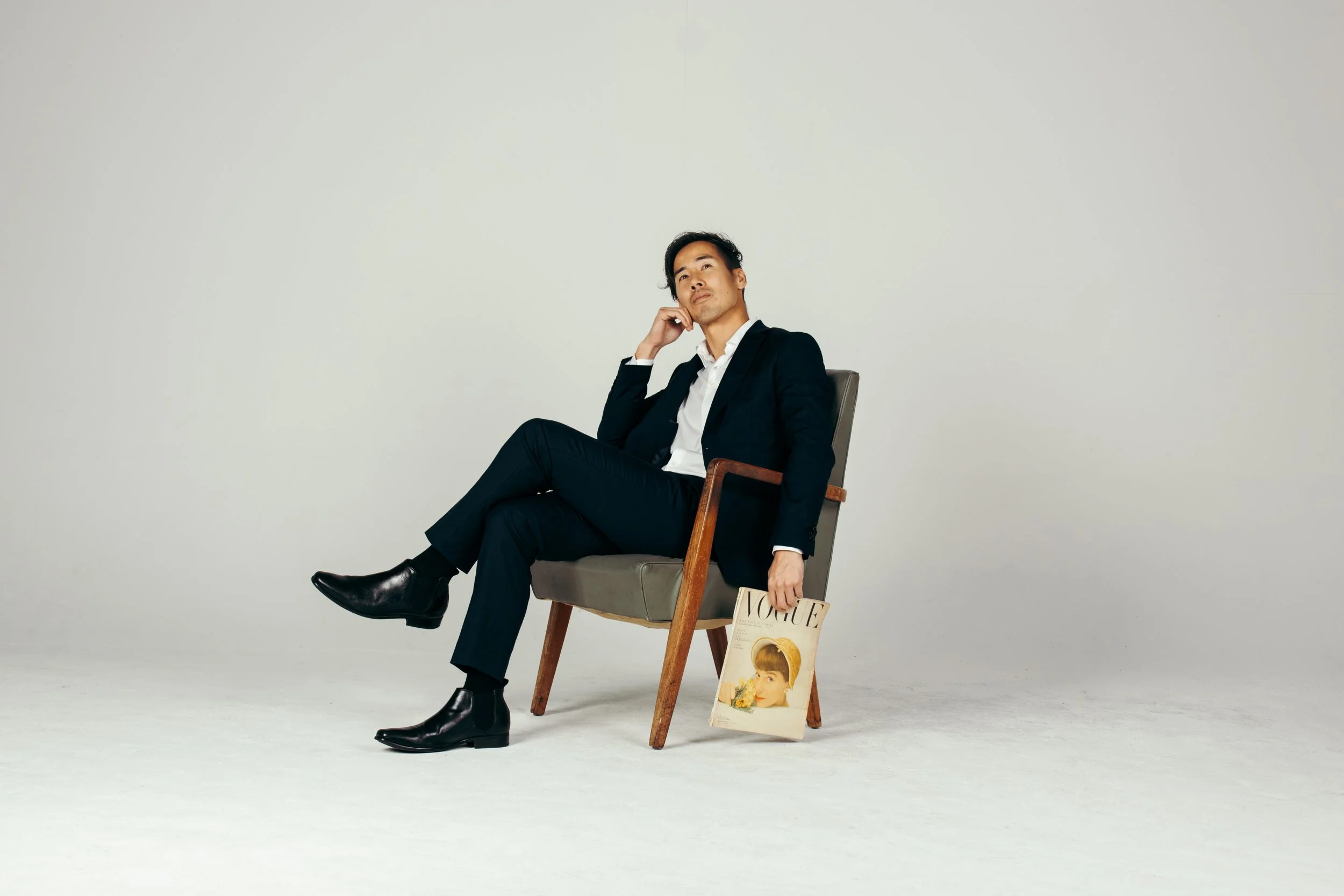 A man in a black suit and white shirt sits on a wooden chair, holding a Vogue magazine and resting his chin on his hand, posed against a plain white background.