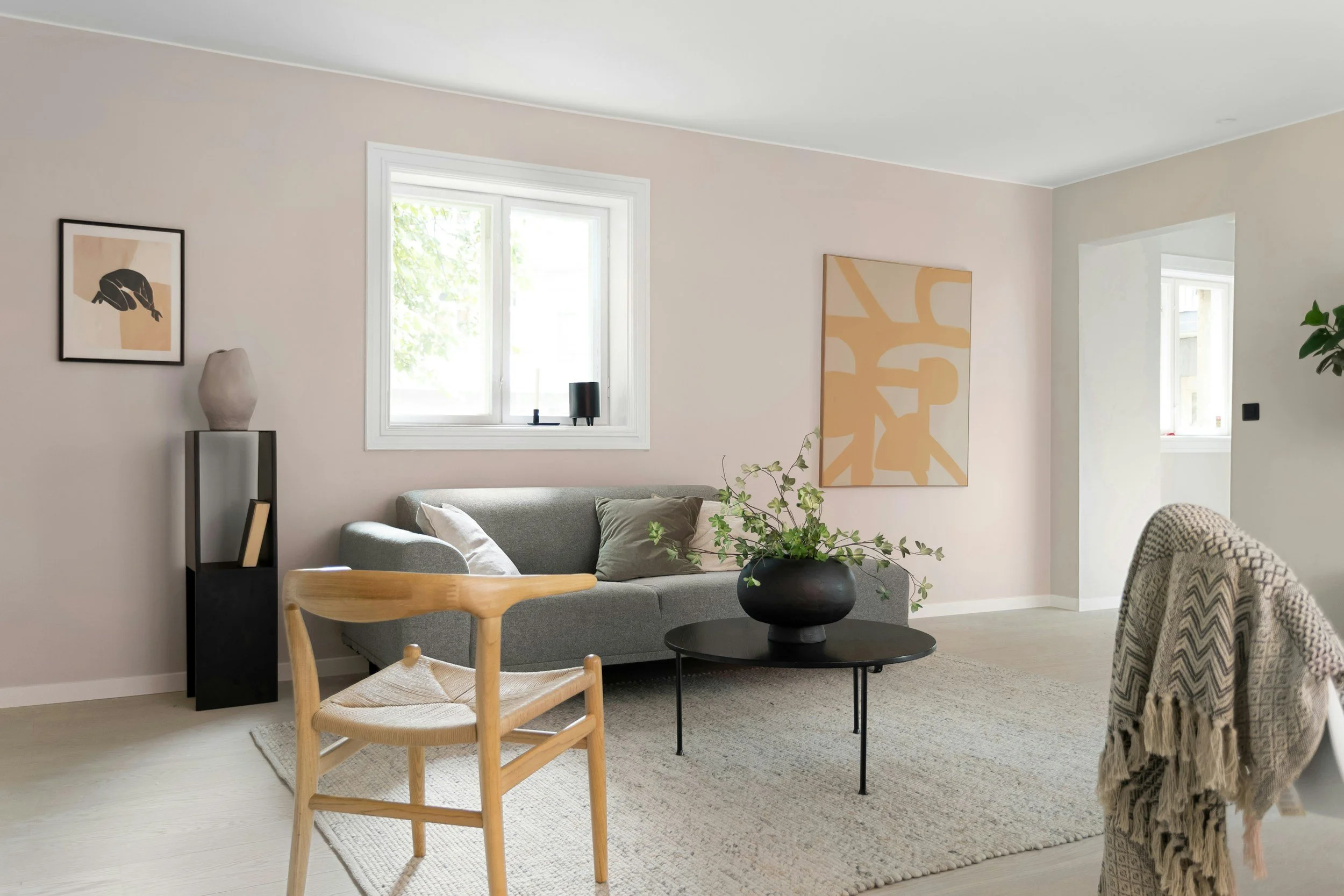 A living room with beige walls, a window with white trim, a gray sofa with cushions, a black round coffee table with a plant, abstract artwork on the wall, a wooden chair, and a beige rug.