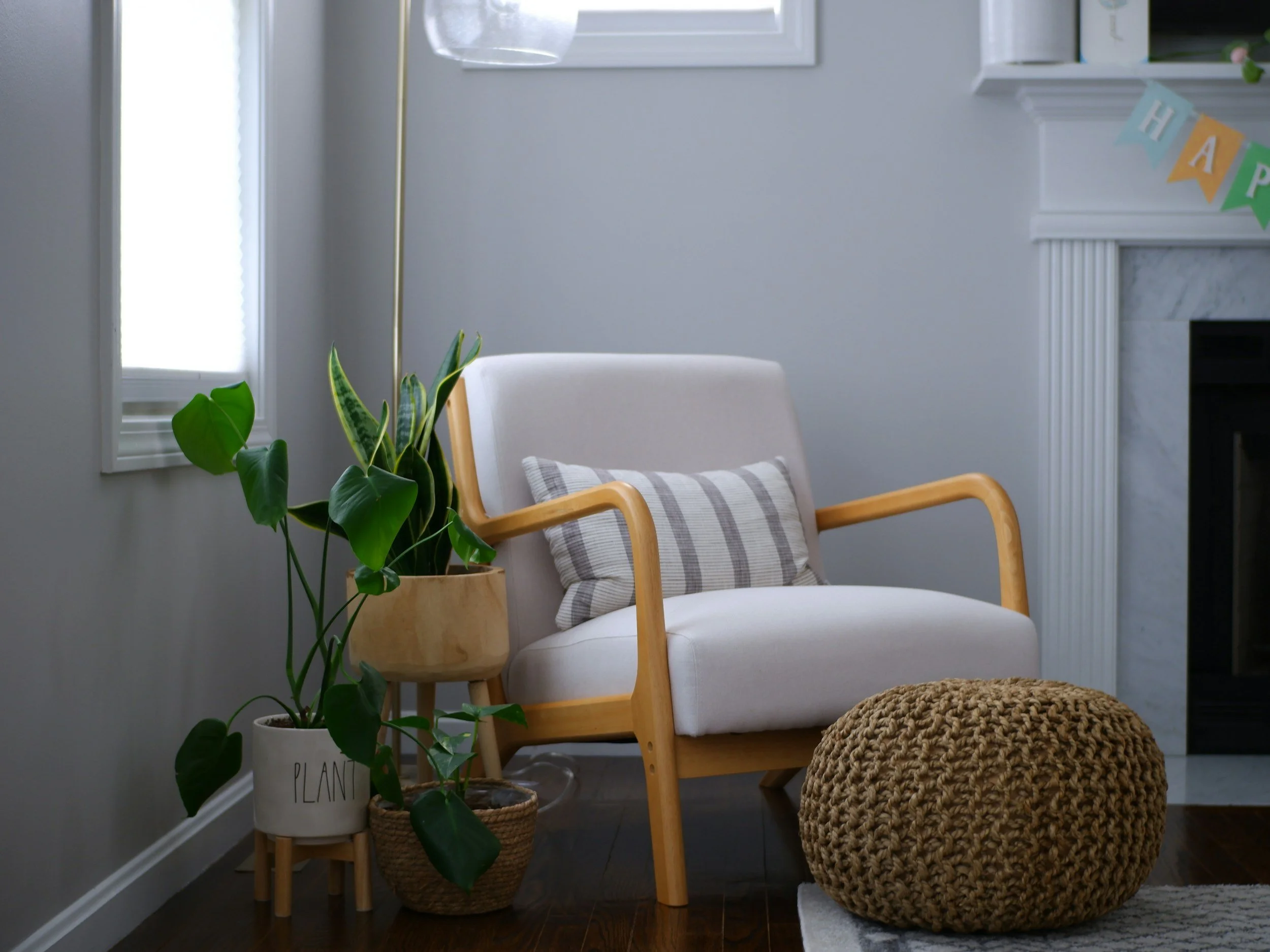 A cozy corner of a room featuring a white armchair with a gray and white striped pillow, a woven pouf in front, a tall potted plant with large green leaves, a small wooden stand, a white wall with a window, and a fireplace with a marble mantel decorated with a colorful 'HAPPY' banner.